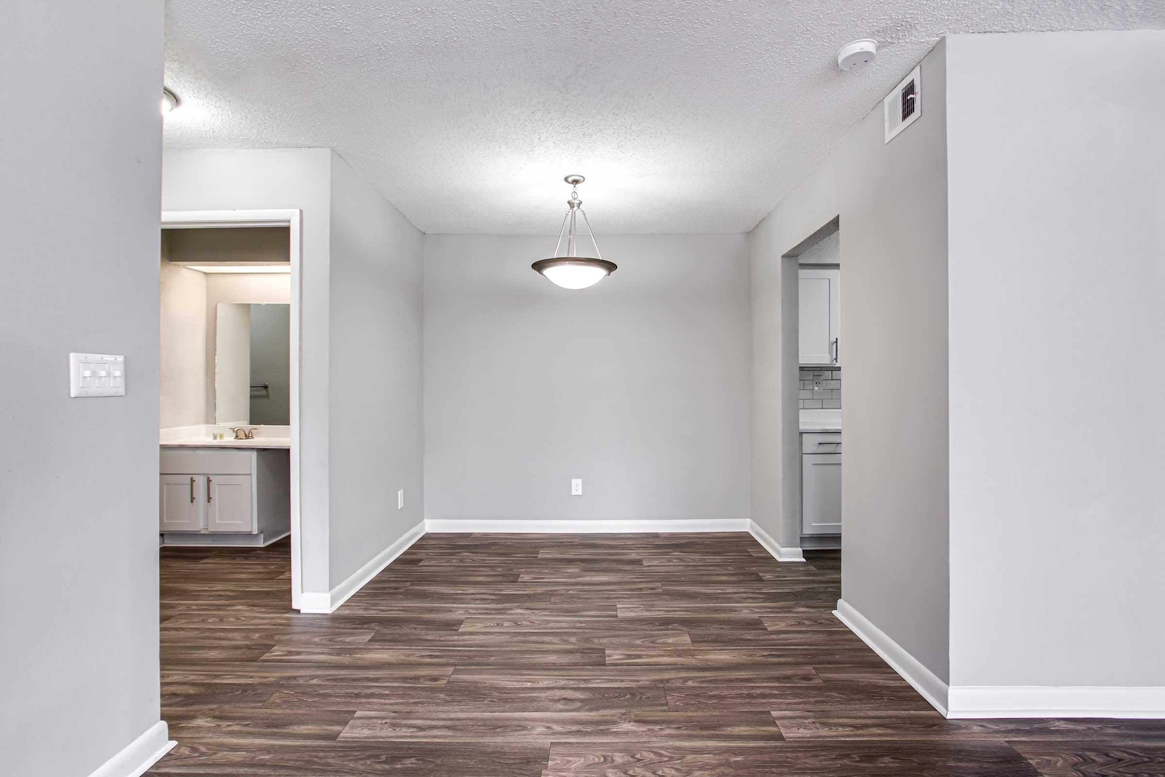 Empty room with light gray walls and dark wood flooring. A ceiling light fixture is hanging in the center. To the left, there is an open doorway leading to a bathroom with a sink visible. To the right, a doorway opens to another room or kitchen area. Overall, the space is well-lit and spacious.
