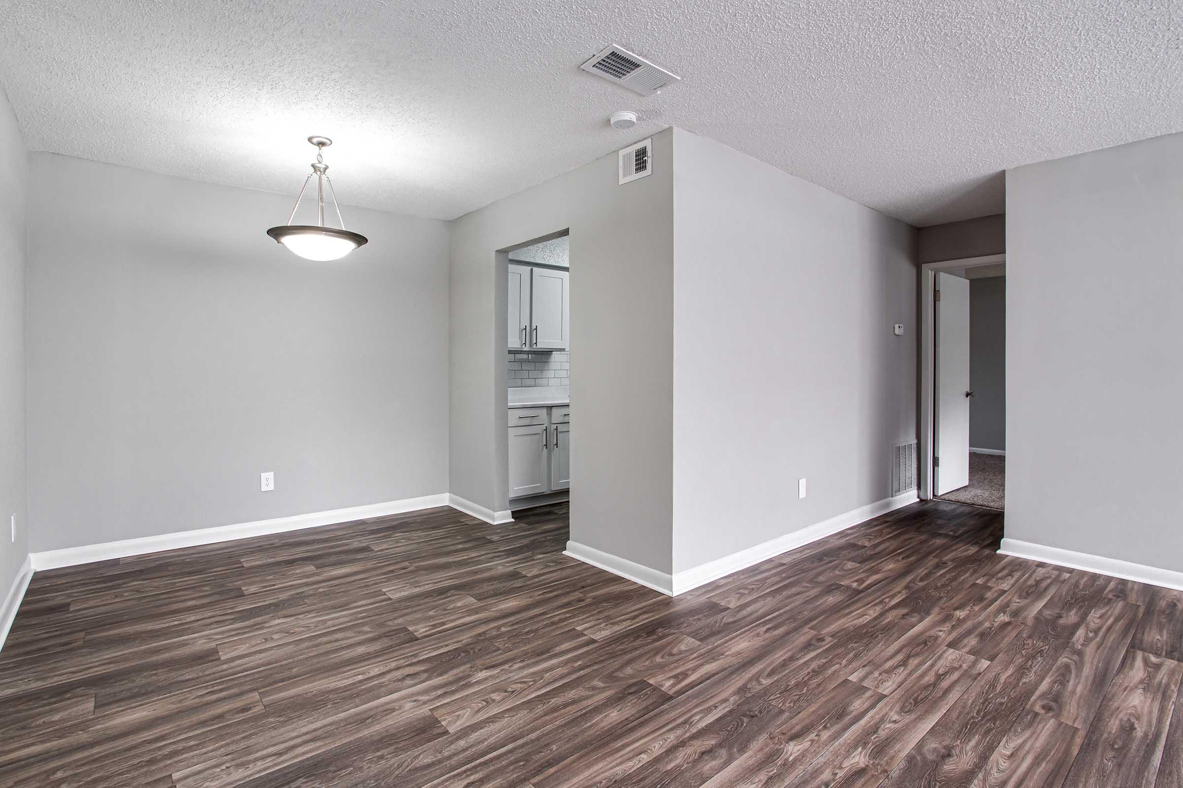 Spacious modern living area with hardwood-like flooring, featuring a light fixture and open layout. A doorway to the right leads to another room, while to the left, a kitchen area is visible through an archway, complete with white cabinetry and a tile backsplash. Walls are painted in a neutral gray tone.