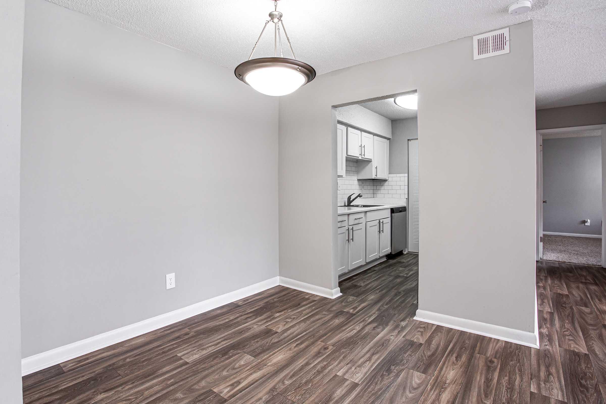 Interior of a modern apartment showcasing a spacious dining area with gray walls and hardwood flooring. A doorway leads to a kitchen with white cabinetry and tiled backsplash. There’s a ceiling light fixture providing illumination, enhancing the overall airy and inviting atmosphere of the space.