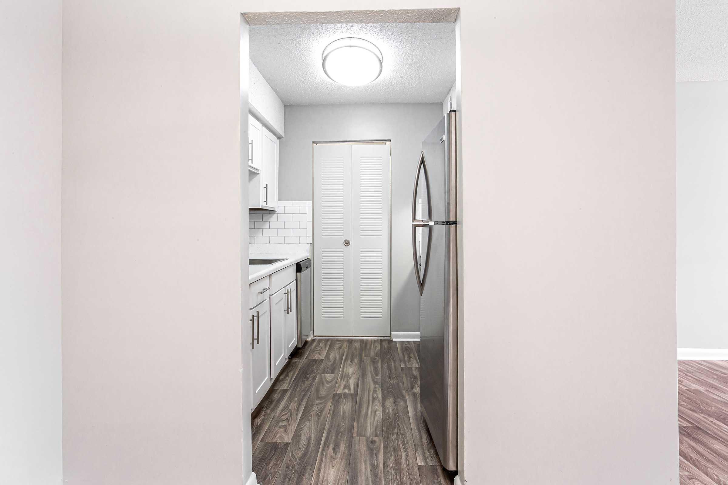 View of a modern kitchen through an arched doorway, featuring gray walls, white cabinets, a stainless steel refrigerator, and a tiled countertop. The floor is a dark wood finish, and there is a door with closed louvered panels on the right side. Soft lighting from a ceiling fixture illuminates the area.