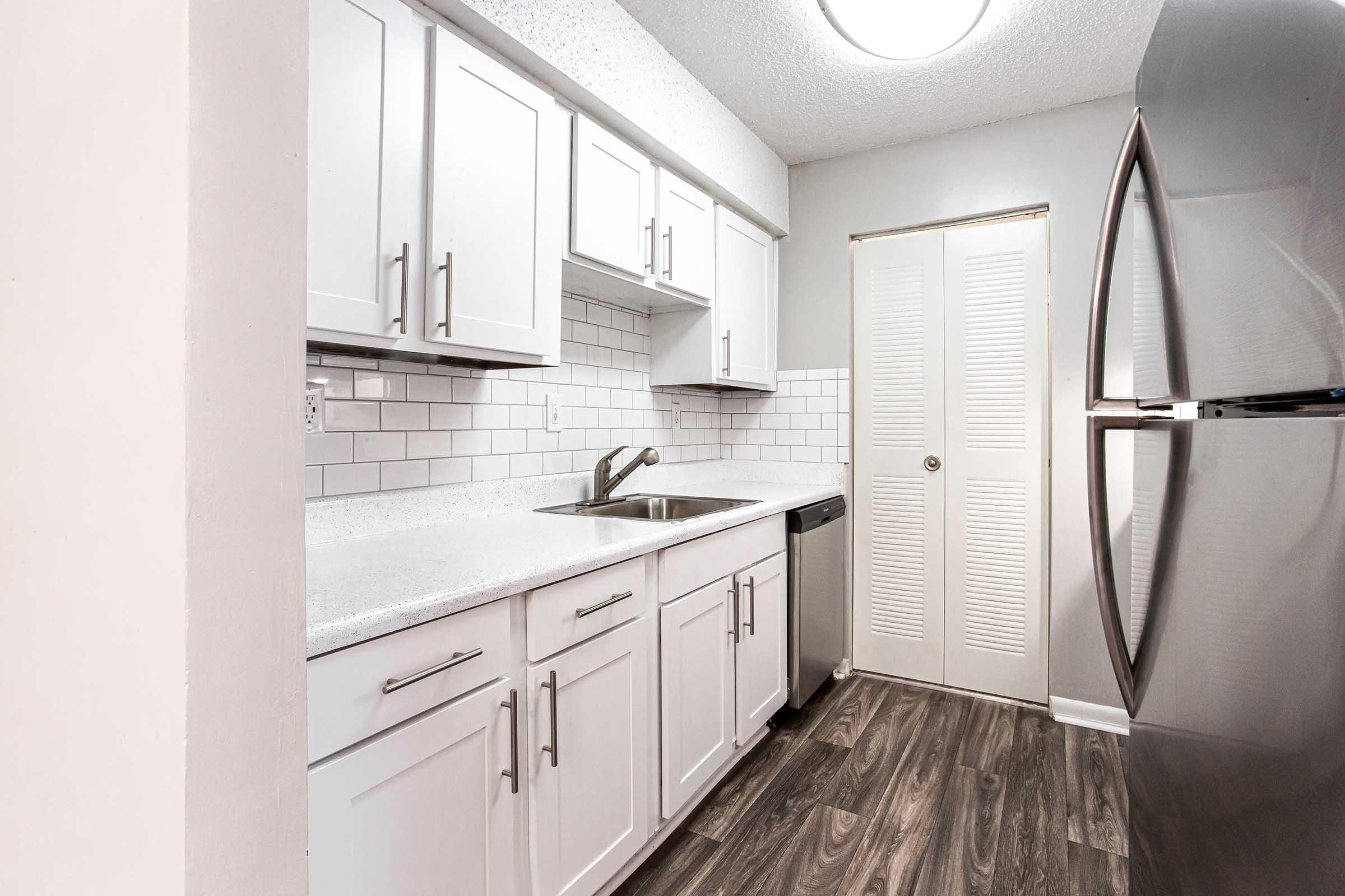 A modern kitchen featuring white cabinets, a gray countertop, and stainless steel appliances. The backsplash consists of light-colored tiles. The flooring is dark wood-like laminate, and there is a closed closet door in the background. Natural light comes from a ceiling fixture, creating a bright atmosphere.
