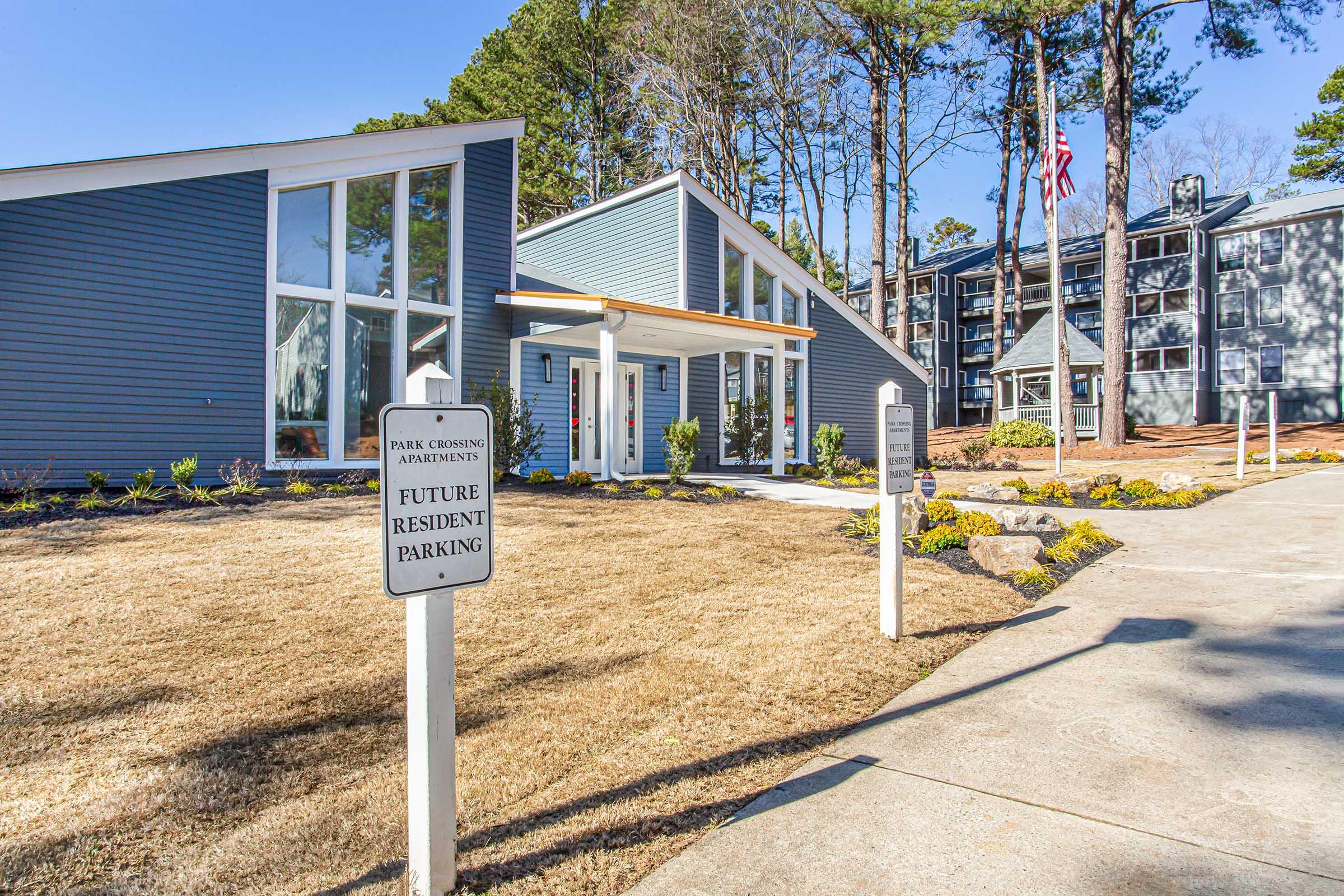 Modern apartment complex entrance featuring a blue building with large windows, a landscaped area with flower beds, and a sign for future resident parking. Flags and trees are visible in the background, creating a welcoming atmosphere.