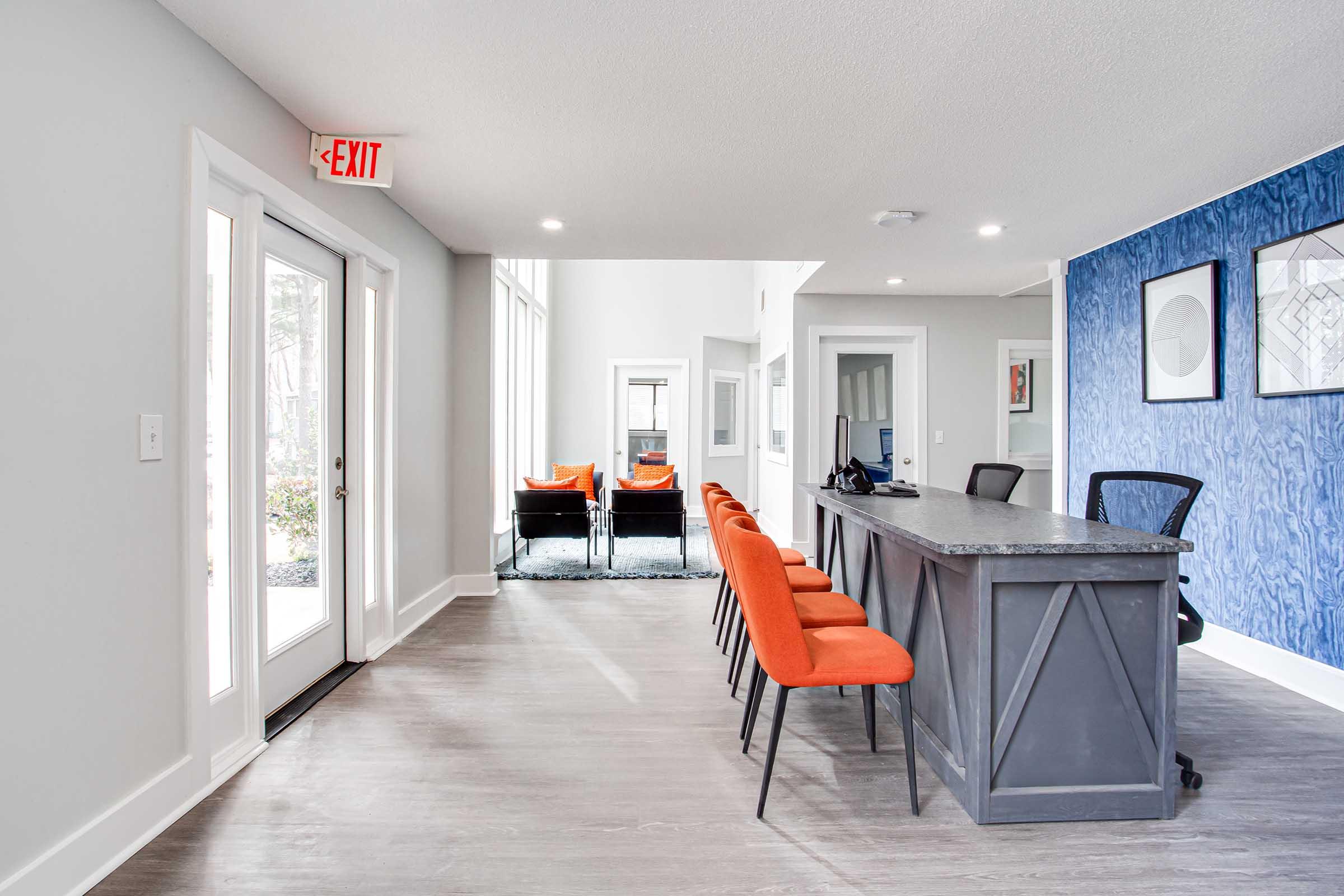 A modern lobby with a gray reception desk and orange stools. Large windows allow natural light, and there are two black chairs on a light carpet in the background. The walls are painted light gray, with one wall featuring a blue textured design. An exit sign is visible above the door.