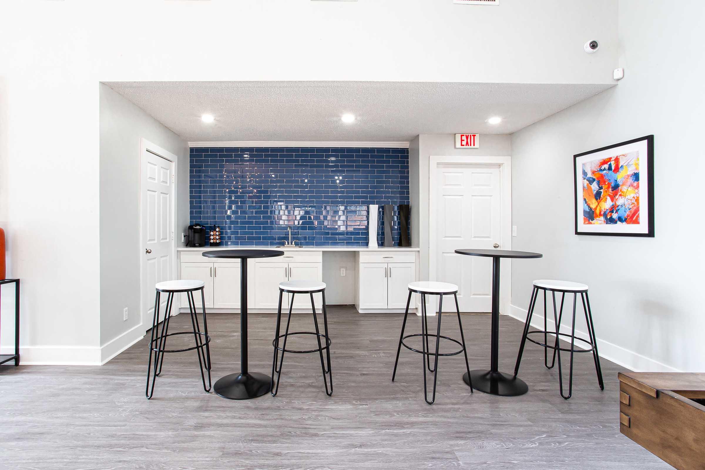 A modern kitchen area featuring a blue tiled backsplash, white cabinetry, and a coffee machine. In the foreground, there are four tall, round tables with black bases and white seats. The flooring is light grey, and there is a colorful artwork piece on the wall.