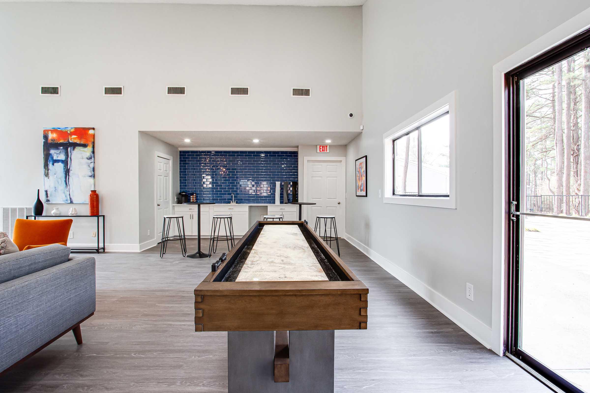 Modern interior of a living space featuring a wooden shuffleboard table in the foreground. The background includes a kitchen area with navy blue tiled walls, bar stools, and a window allowing natural light. The decor includes vibrant artwork and clean lines, creating a stylish and inviting atmosphere.