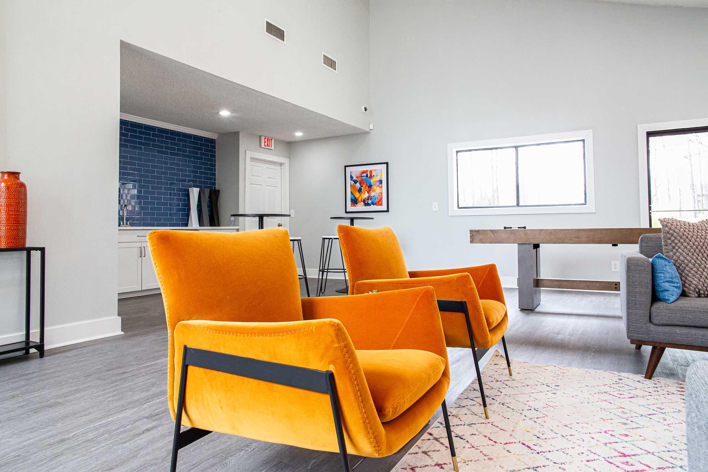 A bright and modern living space featuring two vibrant orange velvet armchairs, a neutral-toned rug, and a large window allowing natural light. In the background, there is a kitchen area with blue tile, a framed artwork, and a wooden table. The decor combines comfort and contemporary design.
