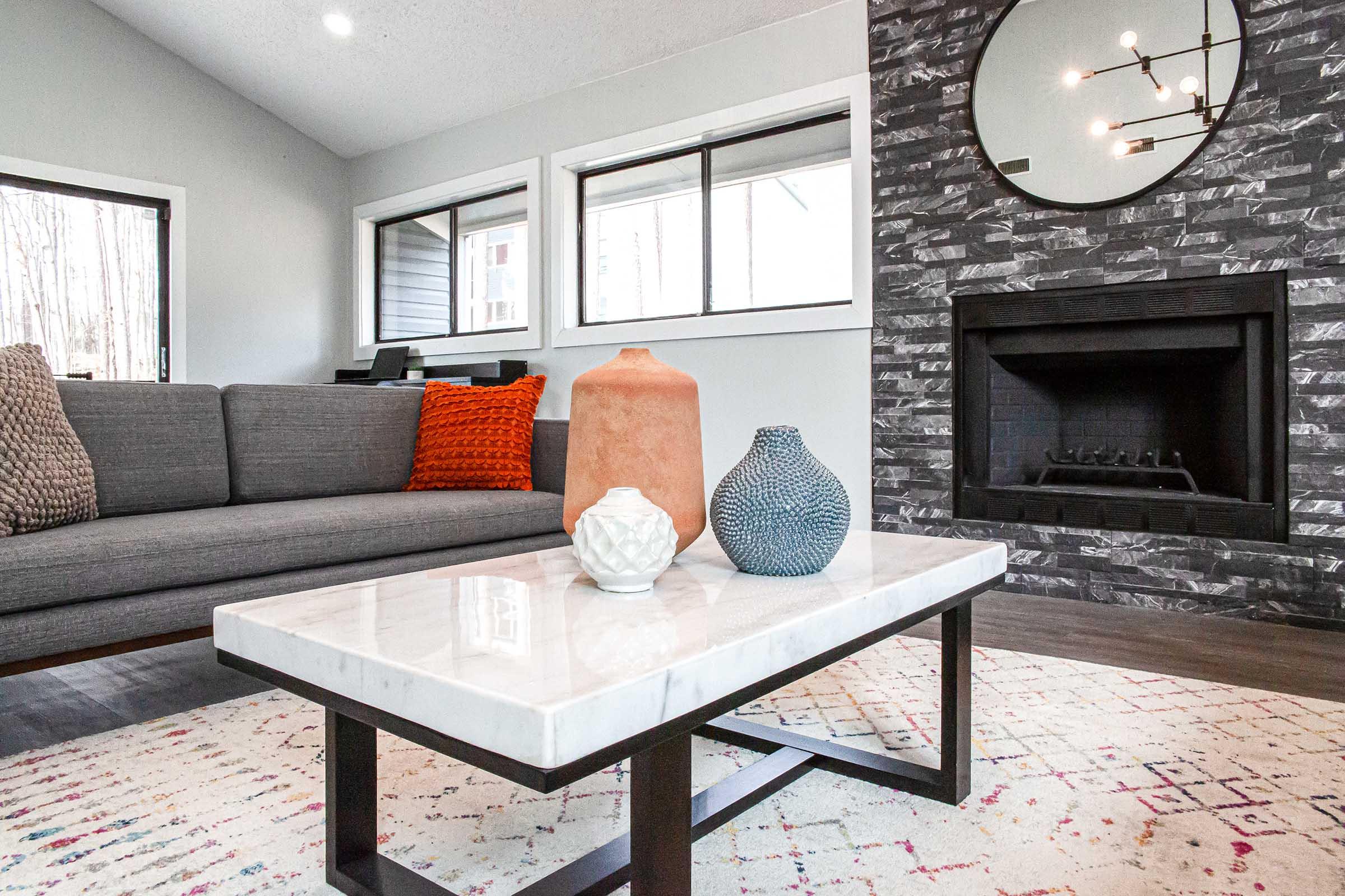 A modern living room featuring a gray sofa with colorful pillows, a coffee table with a marble top and decorative vases, and a stone fireplace. Large windows allow natural light, and a circular mirror is mounted on the wall, enhancing the contemporary design atmosphere.