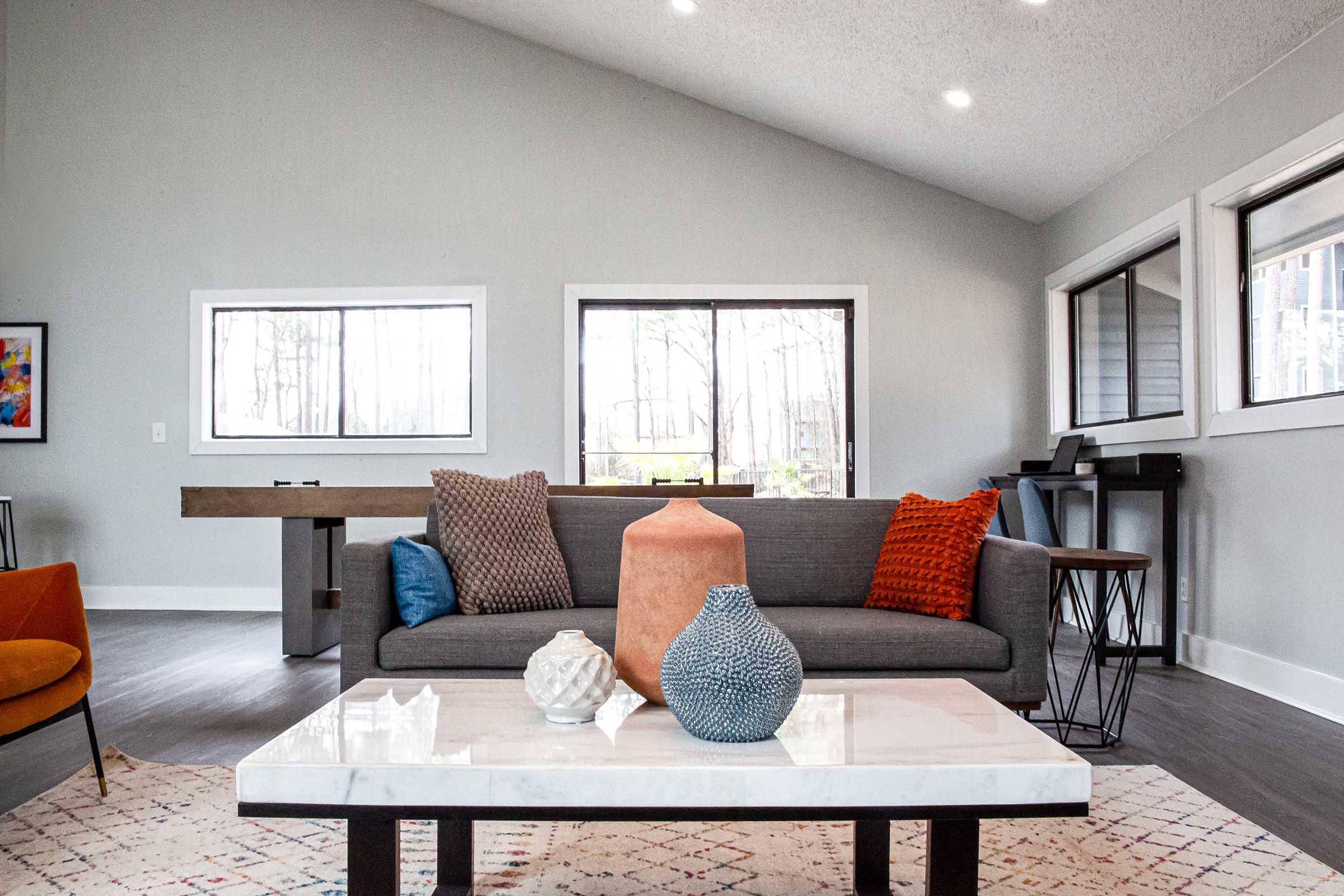 Modern living room featuring a gray sofa with colorful throw pillows, a marble coffee table, decorative vases, and large windows that let in natural light. A wooden bar or console table is visible, along with a cozy orange accent chair, creating a welcoming and stylish atmosphere.