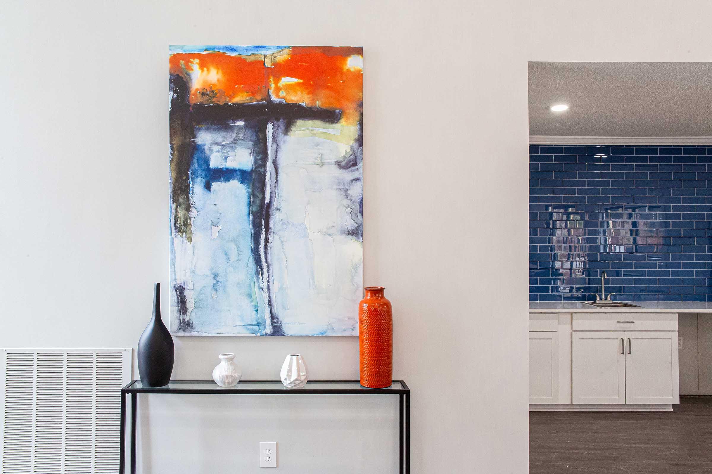 A modern living space featuring an abstract painting with orange and blue tones mounted on a light wall. Below it, a sleek glass console table holds a black vase, a small white vase, and a transparent vase. In the background, a kitchen area with blue tile backsplash and white cabinetry is visible.