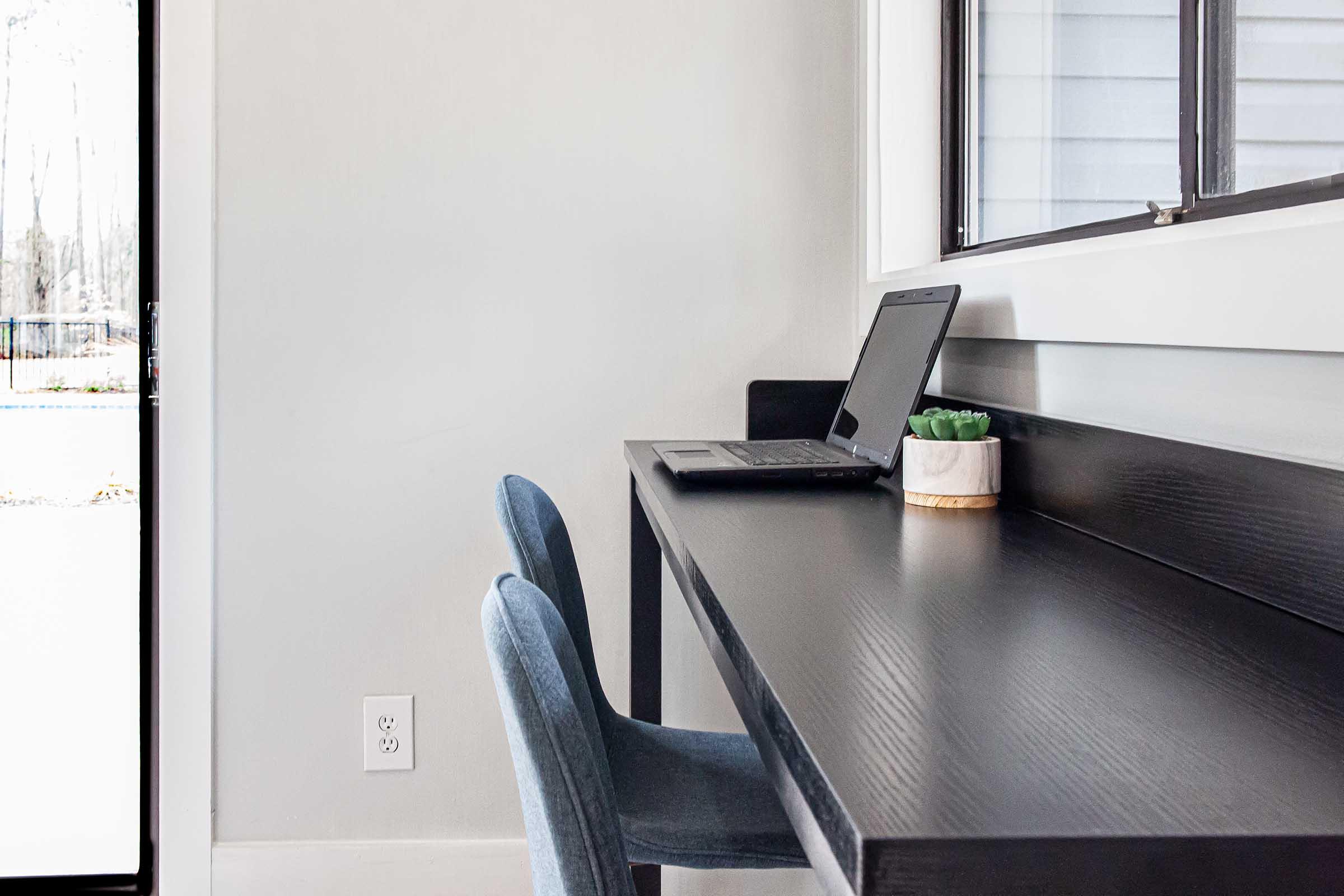 A minimalist workspace featuring a black desk with a laptop, two blue chairs, and a small potted plant. Natural light streams through a nearby window, creating a bright and inviting atmosphere. The wall is painted a light color, enhancing the modern aesthetic of the room.