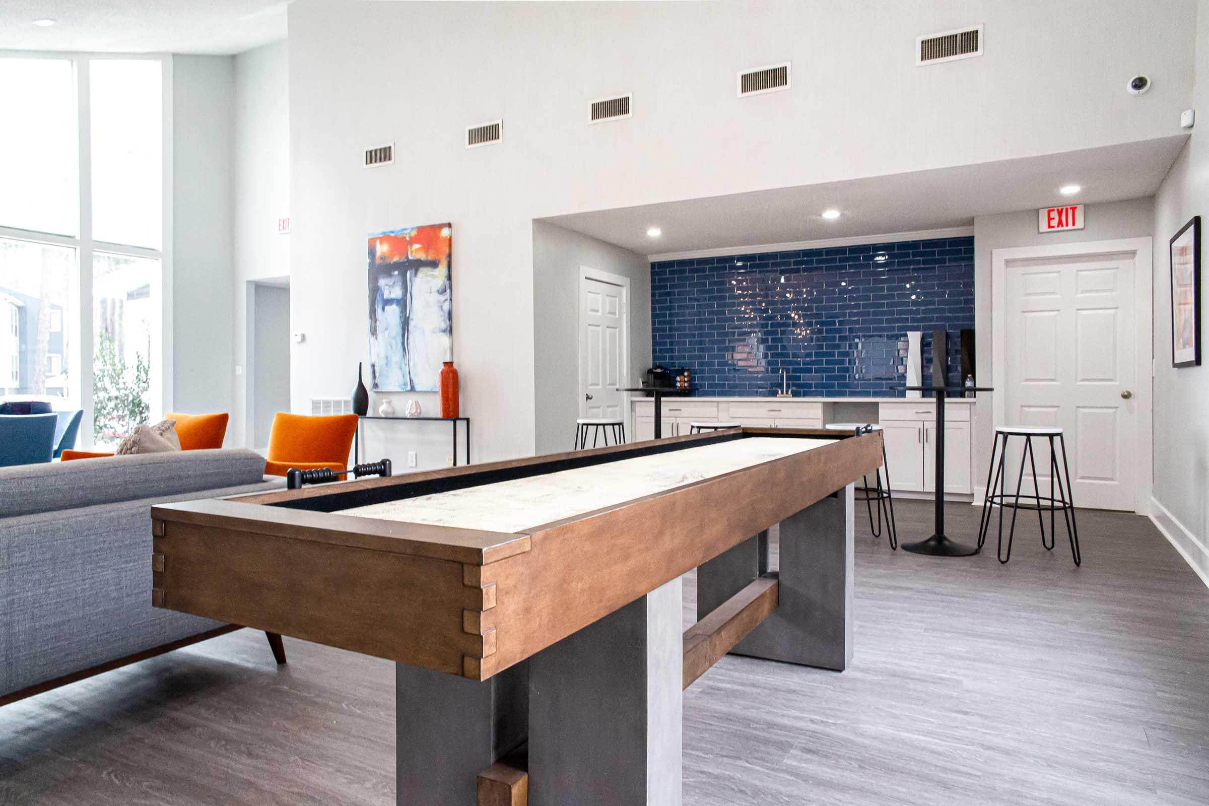 A modern community lounge featuring a pool table with a wood and metal design, gray sofas, and vibrant orange armchairs. The background includes a blue tiled wall, a kitchenette, and tall black stools. Large windows allow natural light to fill the space, enhancing the contemporary atmosphere.