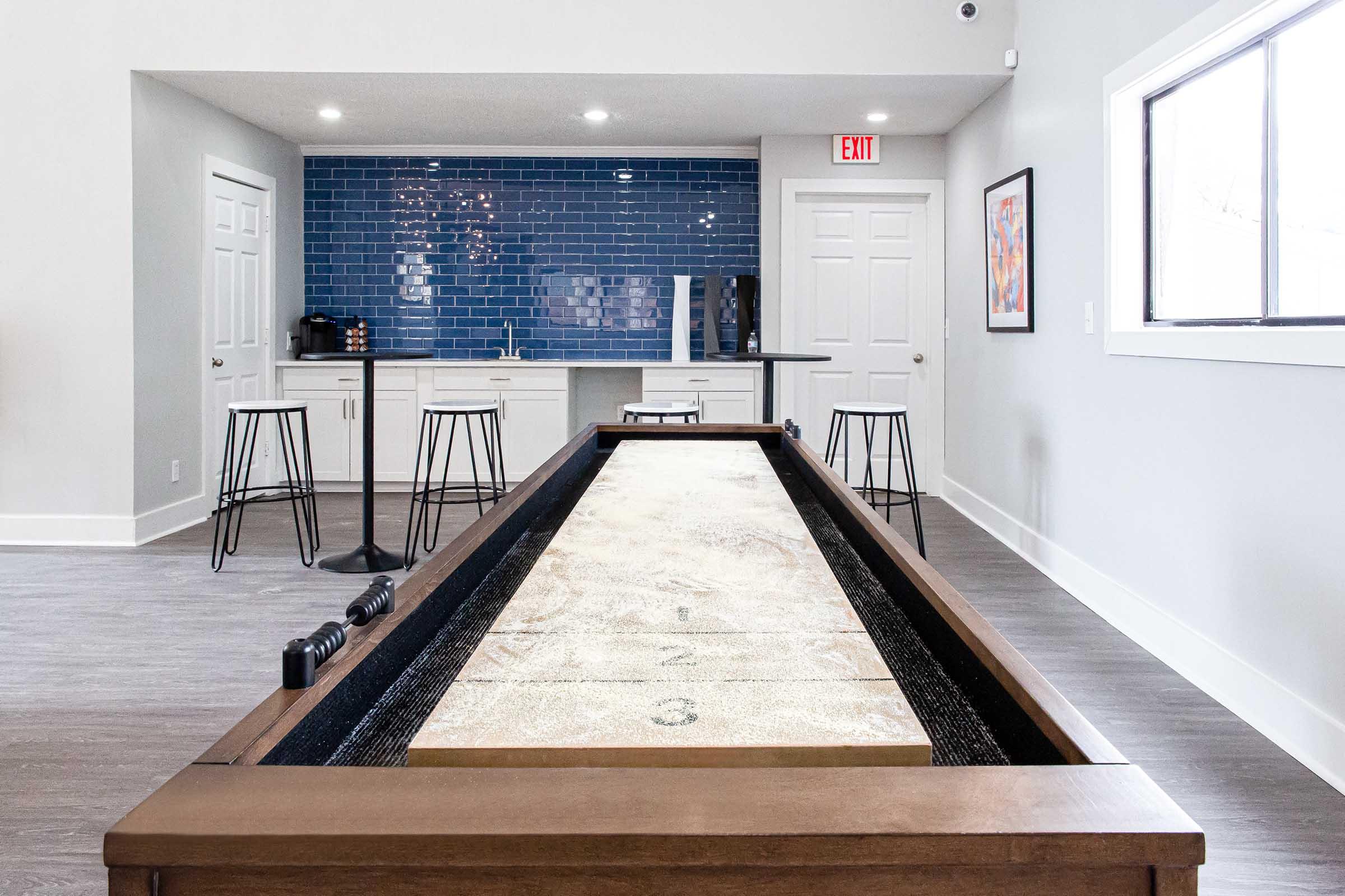 A spacious room featuring a shuffleboard table in the foreground with a wooden finish. In the background, there is a modern kitchen area with blue tiled walls and white cabinets, including bar stools and appliances. The space is well-lit and has a contemporary design.