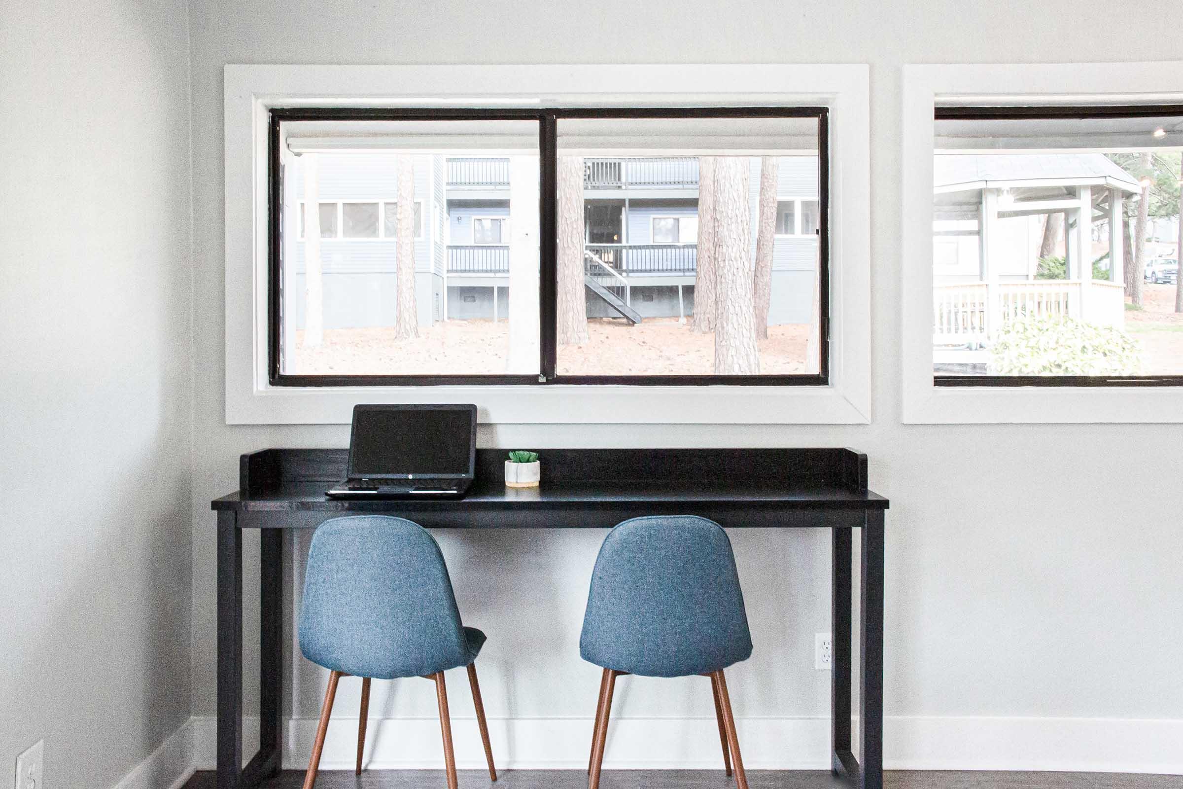 A minimalist workspace featuring a black desk with a laptop and a small potted plant. Two blue chairs are positioned at the desk, facing large windows that provide a view of an exterior building and trees. The interior has light gray walls and a simple, clean aesthetic.