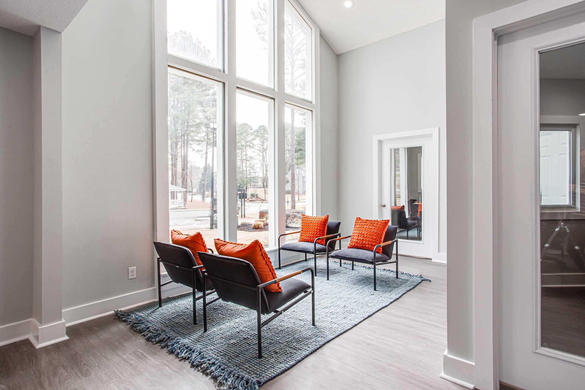 A modern interior space featuring large windows that allow natural light, with a stylish seating arrangement consisting of four black chairs with orange cushions on a textured rug. The walls are painted in soft gray, and there's a mirror reflecting the room, enhancing the open feel.