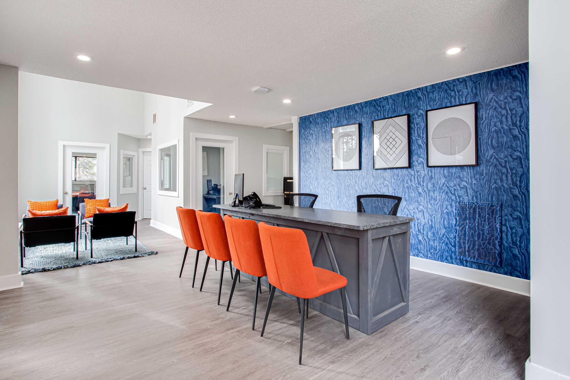 A modern lobby with a gray reception desk and vibrant orange chairs. The wall features a blue design with three framed artworks. Bright lighting illuminates the space, complemented by a cozy seating area with black chairs and orange accent pillows.