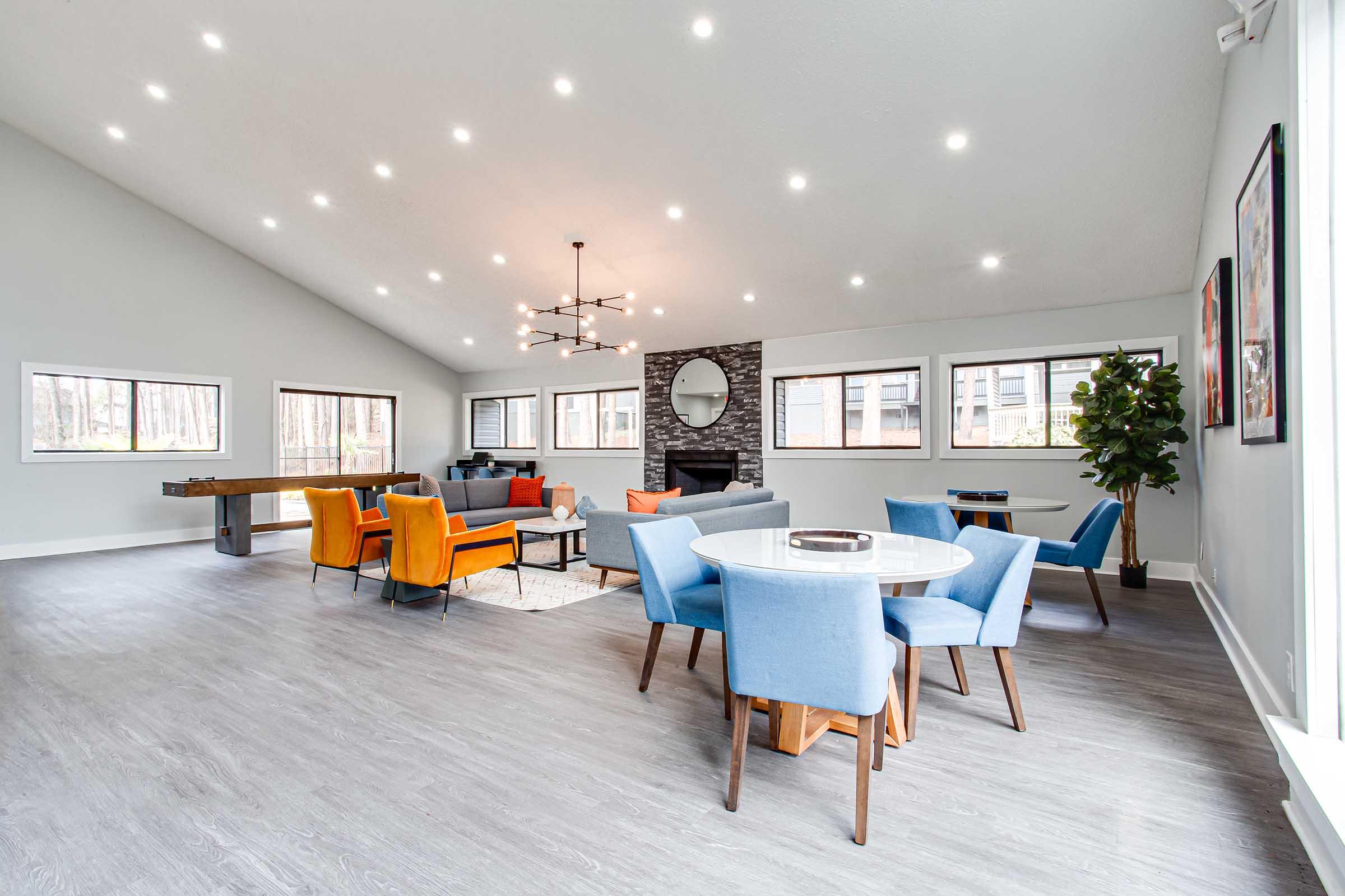 A spacious, modern living area with high ceilings and plenty of natural light. It features a circular dining table surrounded by blue chairs, a cozy seating area with bright orange and blue chairs, a stone fireplace, and large windows showcasing a view of trees outside.