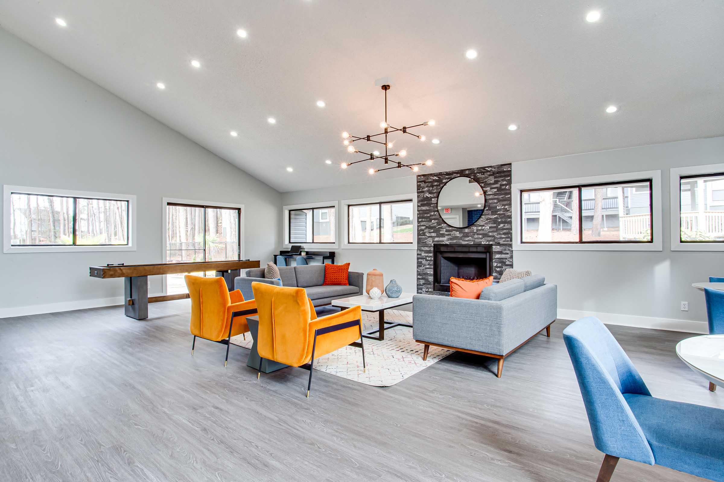 A modern living room featuring a high ceiling, large windows, and stylish decor. The space includes a gray sofa and orange accent chairs, a coffee table, and a fireplace with a circular mirror above it. A dining area with a table is visible in the background, complemented by a neutral-toned area rug.