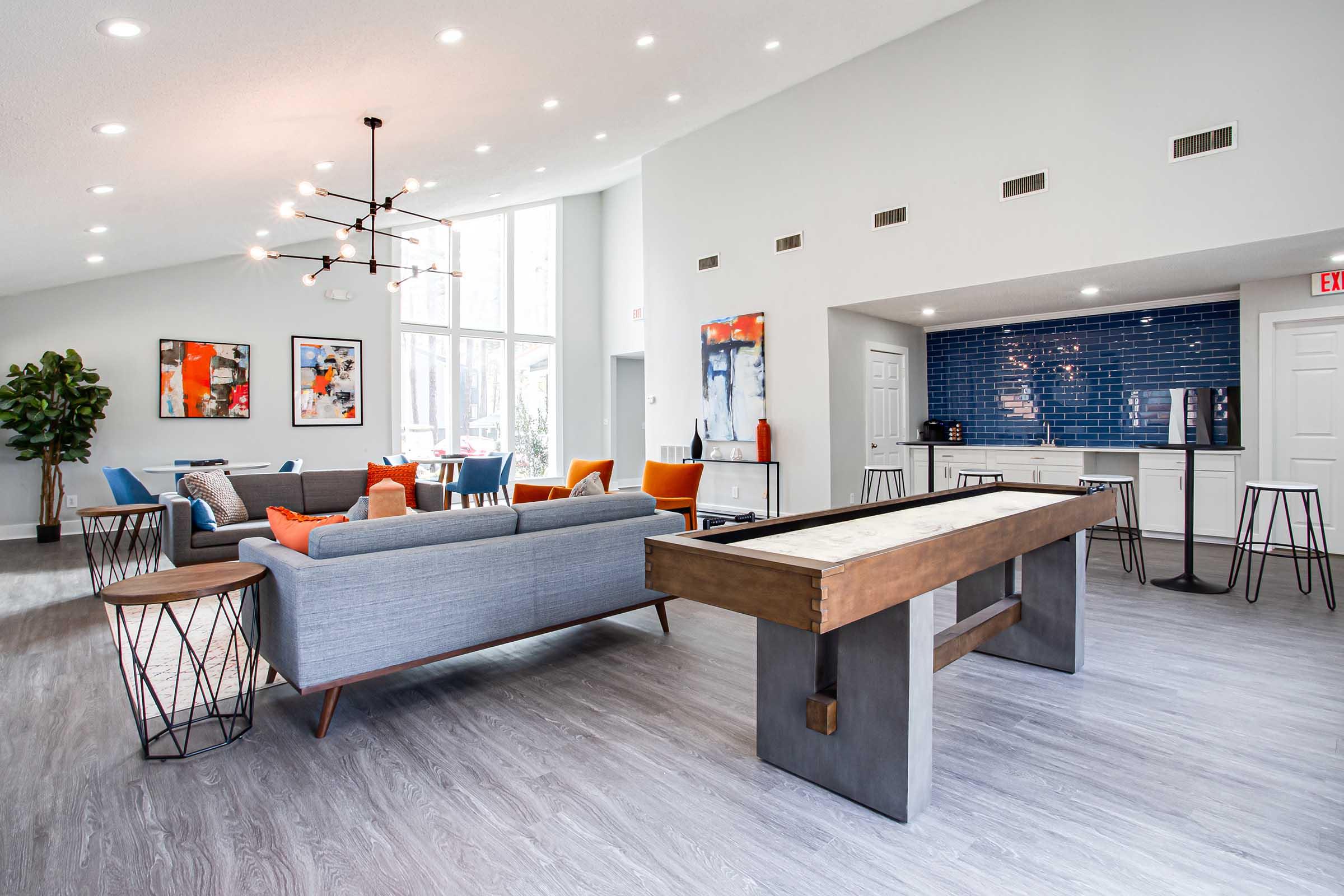 Modern lounge area featuring a gray sofa with colorful pillows, a wooden pool table, and a sleek kitchen space with blue tiled backsplash. Large windows provide natural light, and art pieces adorn the walls, creating an inviting atmosphere.