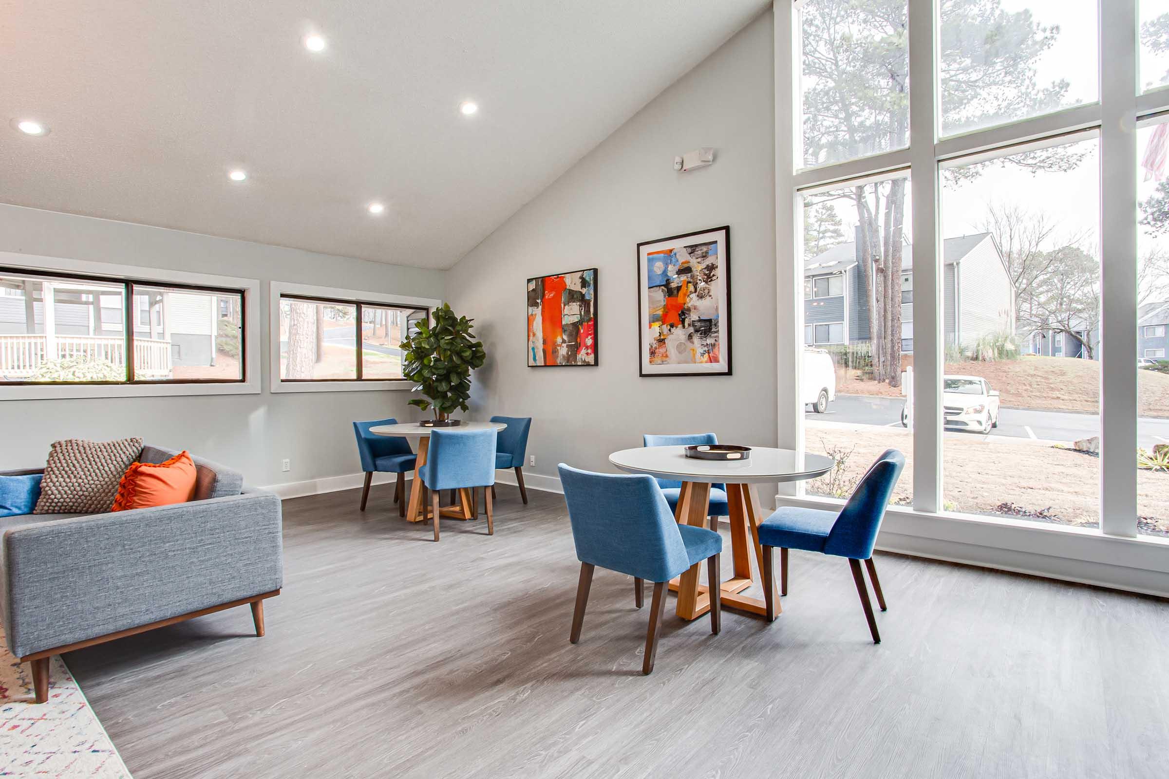 A modern lounge area with large windows allowing natural light. It features a gray sofa with patterned cushions, two small round tables surrounded by blue chairs, and decorative artwork on the walls. A potted plant adds a touch of greenery, and the space is clean and inviting.
