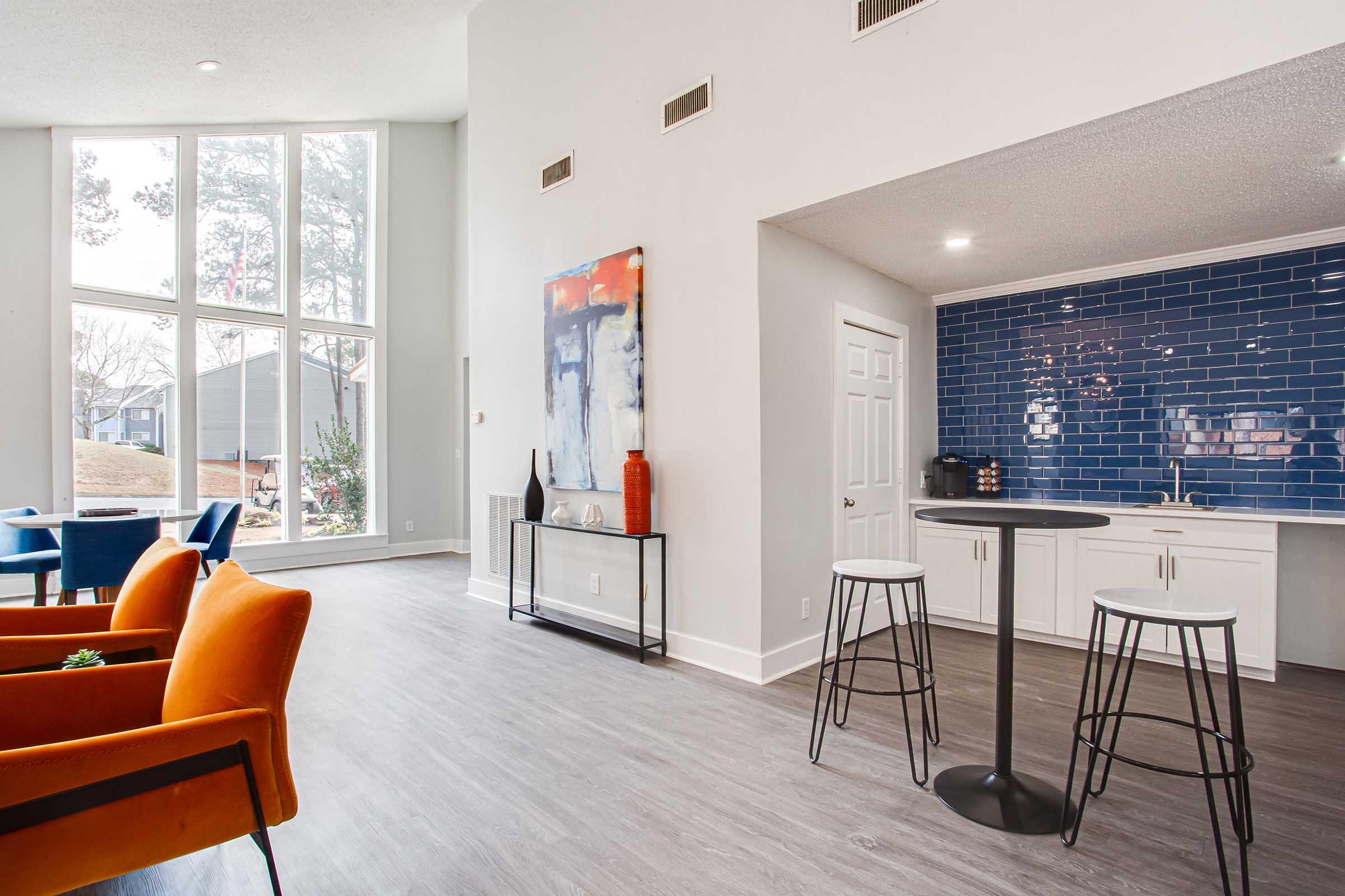 Modern interior of a residential space featuring large windows that allow natural light, a kitchenette with blue tile backsplash, a round black table with two stools, and two orange armchairs. The decor includes abstract art and potted plants, creating a stylish and inviting atmosphere.