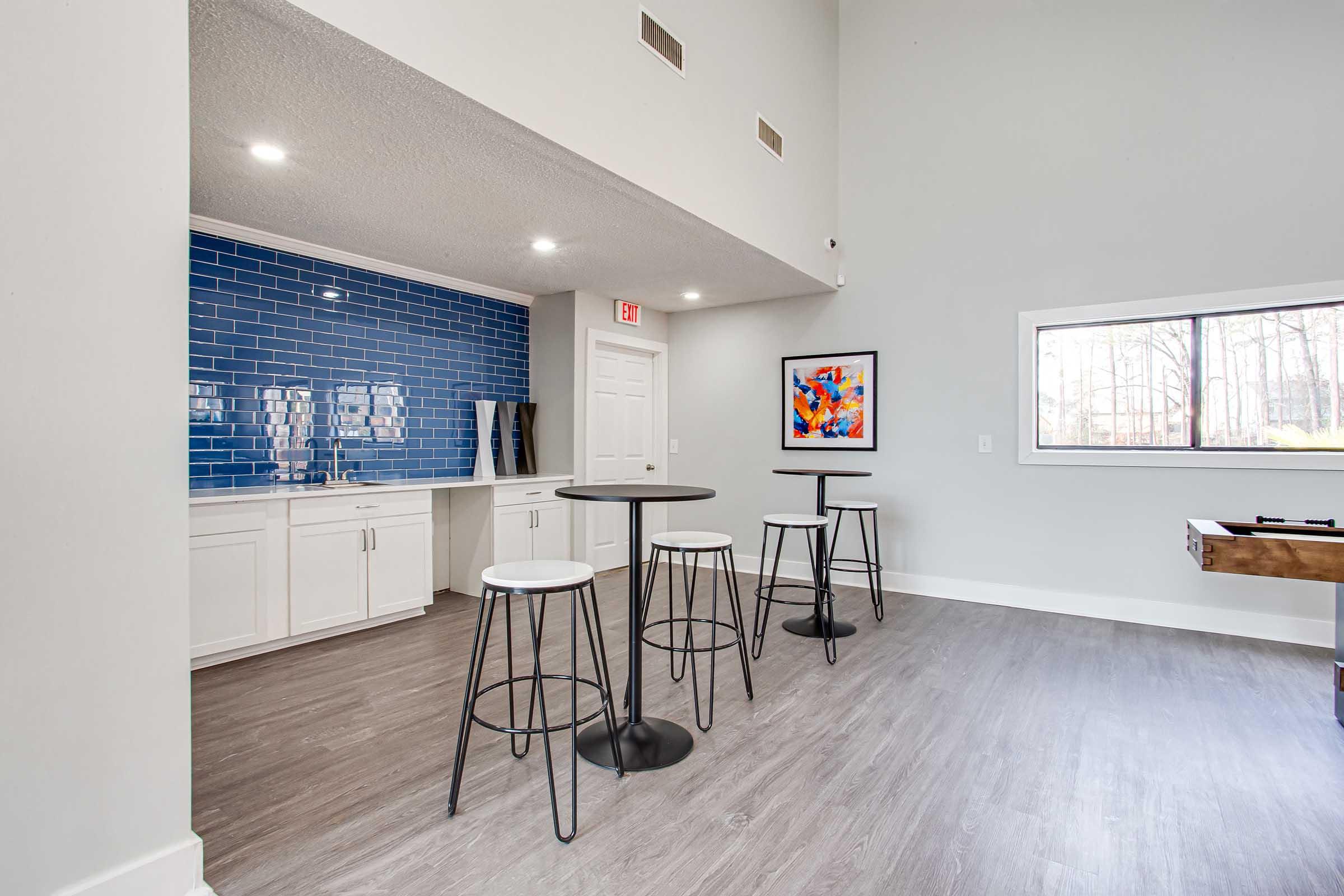 A modern, spacious interior of a lounge area featuring a blue tiled wall, a white countertop, and several tall tables with black stools. A framed piece of artwork is on the wall, and there is a window showing a view of trees outside. The floor is light-colored wood laminate.