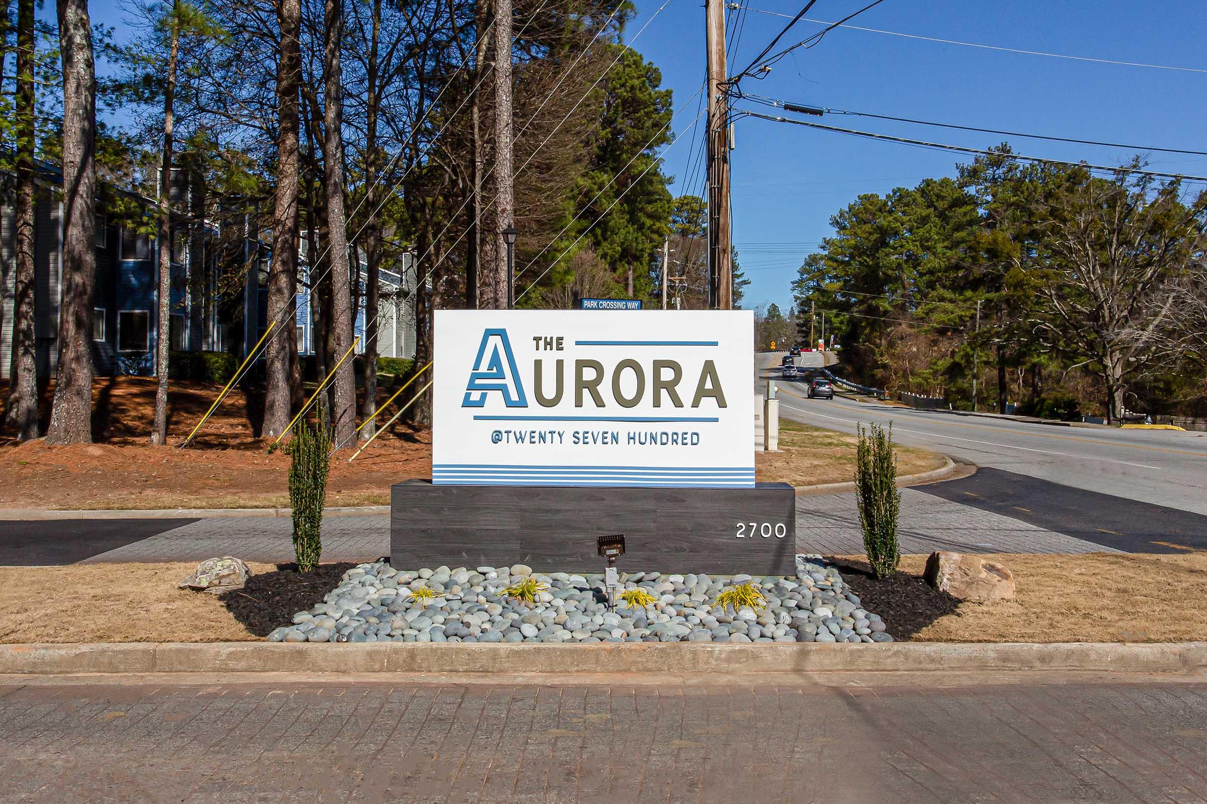 Sign for "The Aurora @ Twenty Seven Hundred" located at 2700, surrounded by landscaping with small rocks and bushes, next to a paved road with trees and power lines in the background.