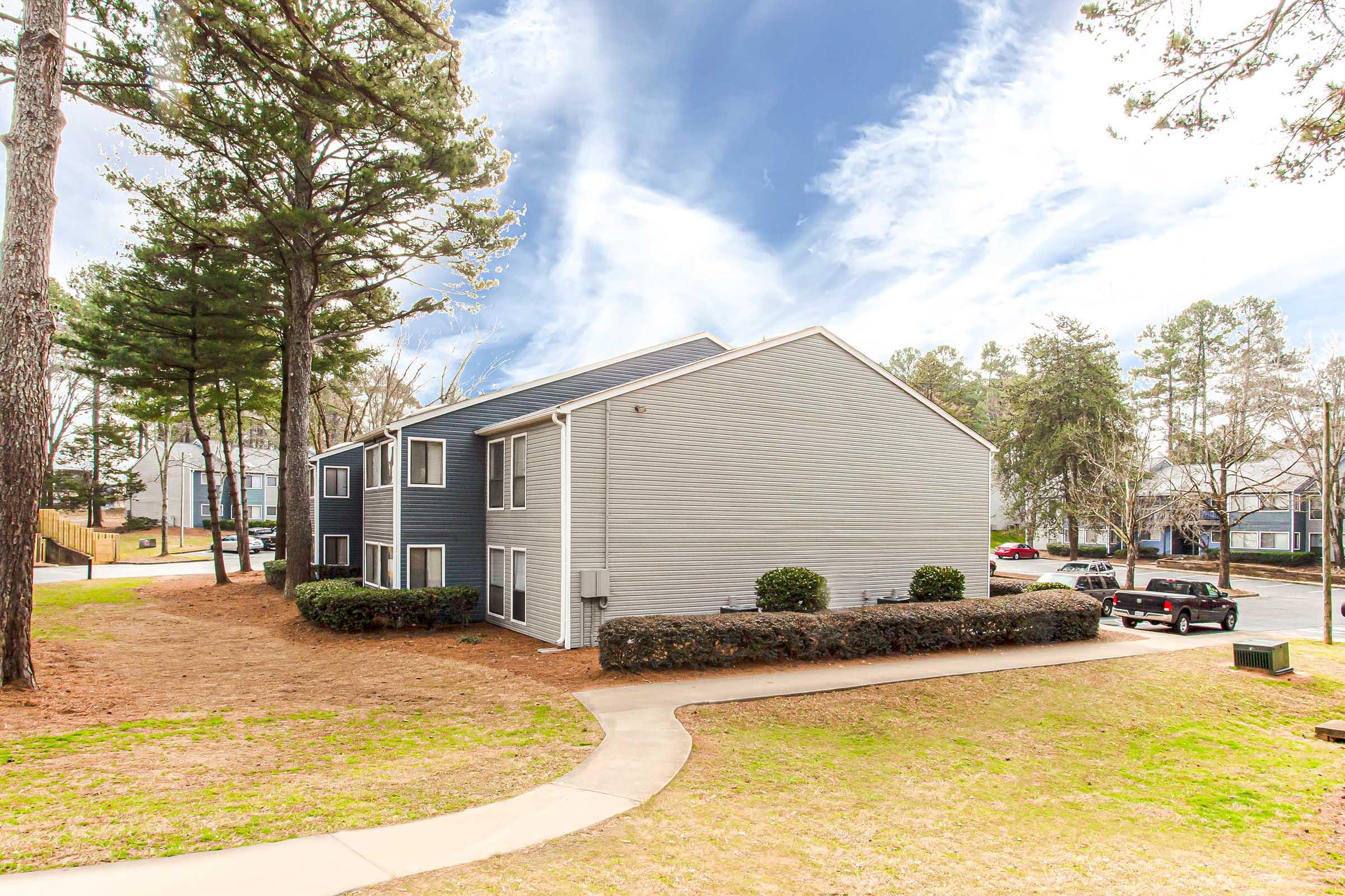 A two-story apartment building with gray siding is surrounded by well-maintained greenery and pine trees. A paved walkway curves in the foreground, leading to the building, and a parked truck is visible nearby on the street. The sky is clear with wispy clouds.
