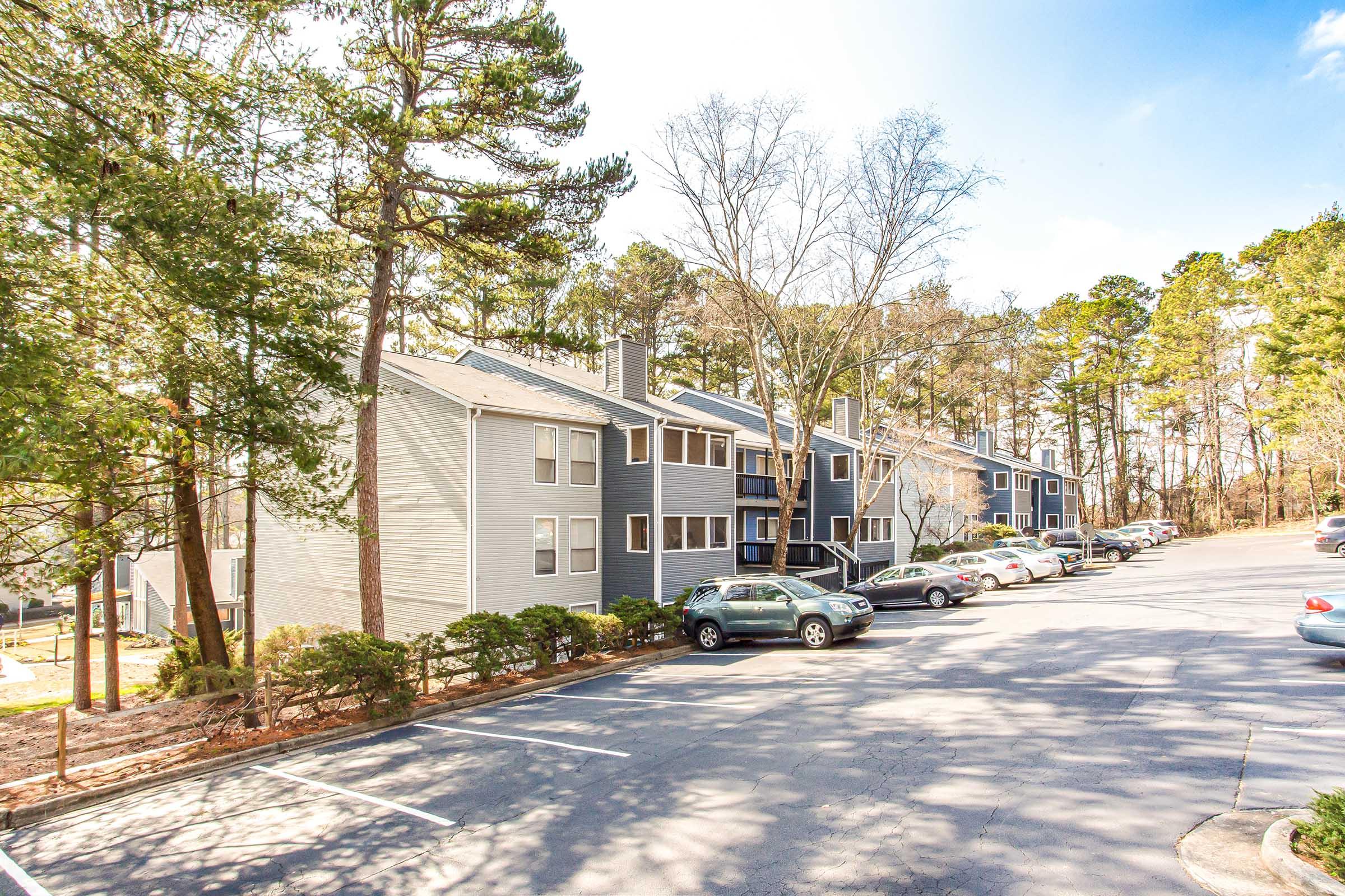 A residential area featuring two-story apartment buildings surrounded by trees. Cars are parked along a paved driveway, with green foliage in the background and a clear blue sky above. The setting appears tranquil and well-maintained, suitable for a suburban living environment.