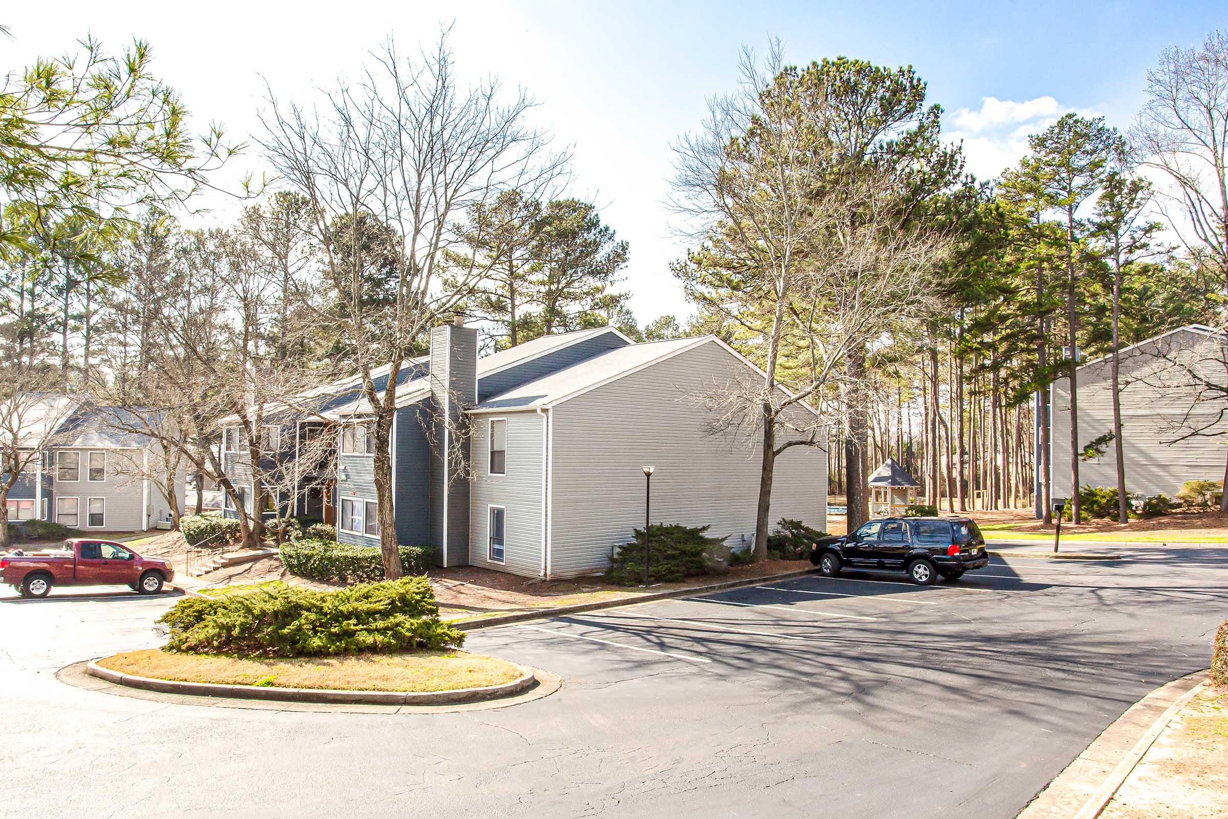 A residential area featuring two multistory apartment buildings amidst trees. The scene includes a circular driveway with parked cars, landscaped bushes, and a clear blue sky with a few clouds in the background.