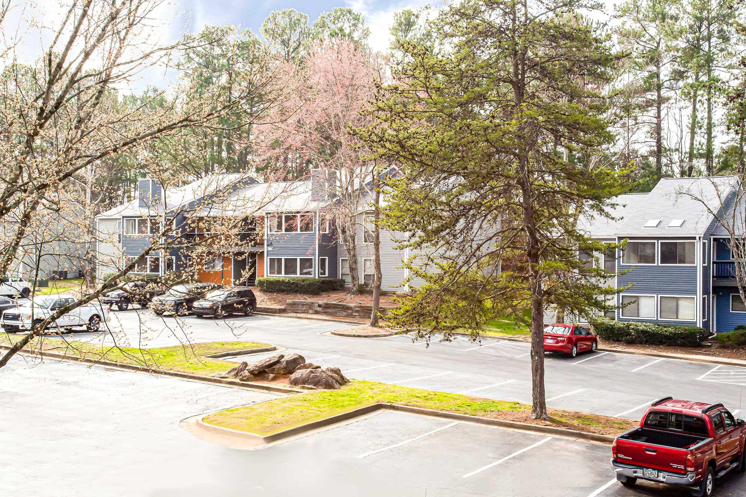 A view of a parking lot outside residential buildings surrounded by trees. Several cars are parked, including red and black vehicles. The scene is partially shaded by trees, with a few scattered clouds in the sky. Springtime foliage is visible, creating a pleasant, suburban atmosphere.