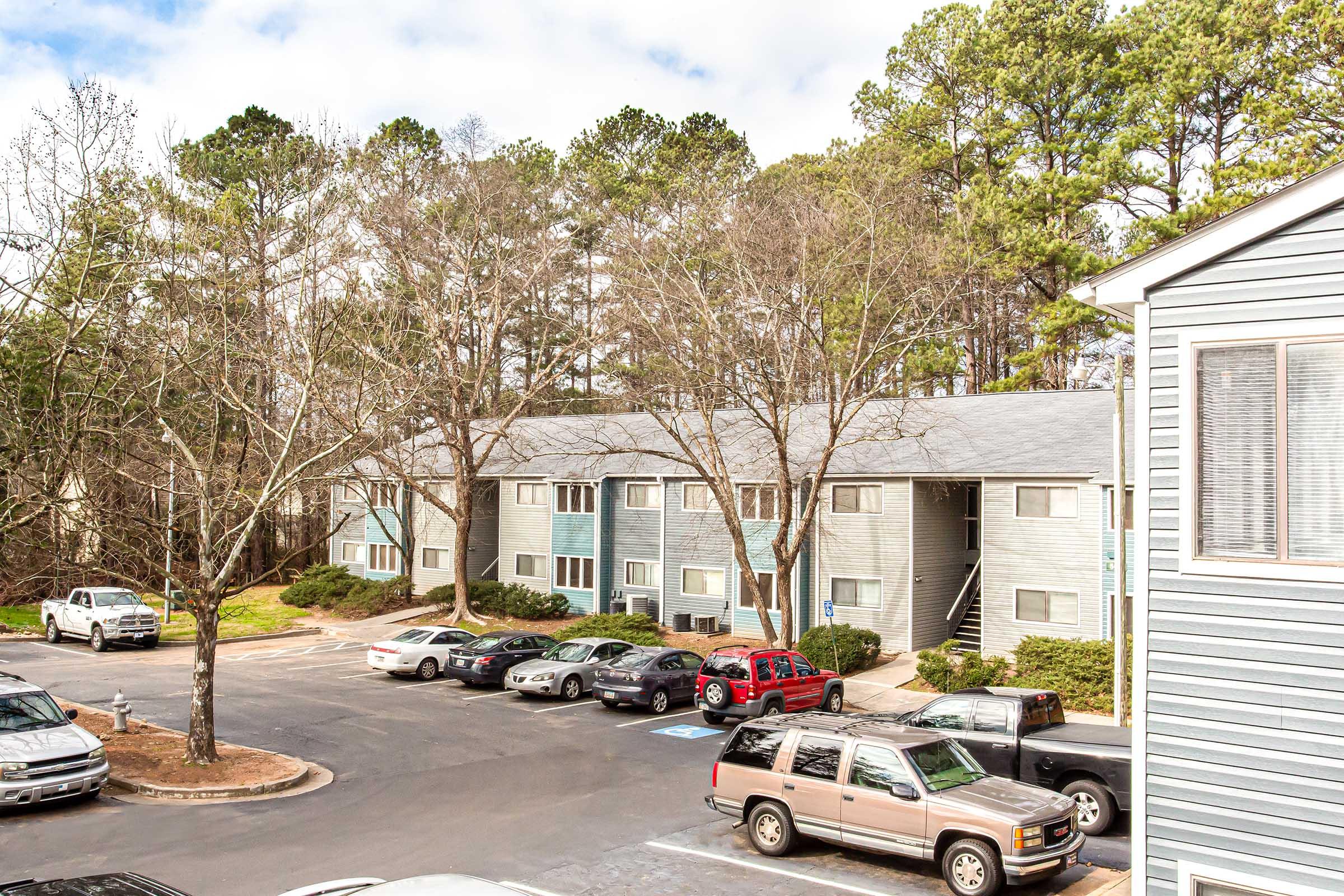 A view of an apartment complex surrounded by trees, featuring multiple two-story buildings. Cars are parked in a lot in the foreground. The scene is partly cloudy, indicating a typical day.