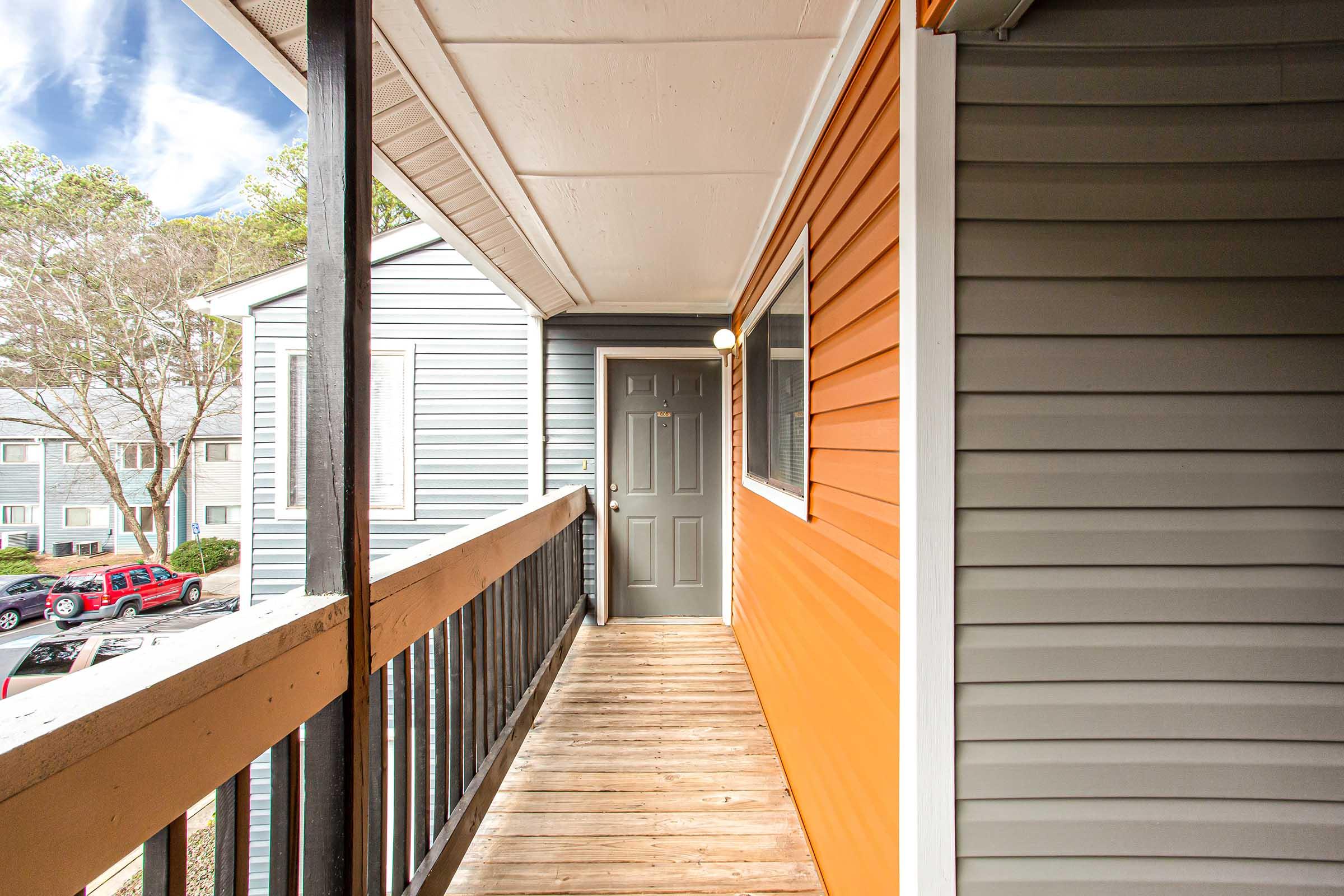 A well-lit corridor featuring wooden flooring and a mix of orange and gray siding on the walls. A gray door is visible at the end of the hallway, with a light fixture above. In the background, there are additional buildings and parked cars visible outside.