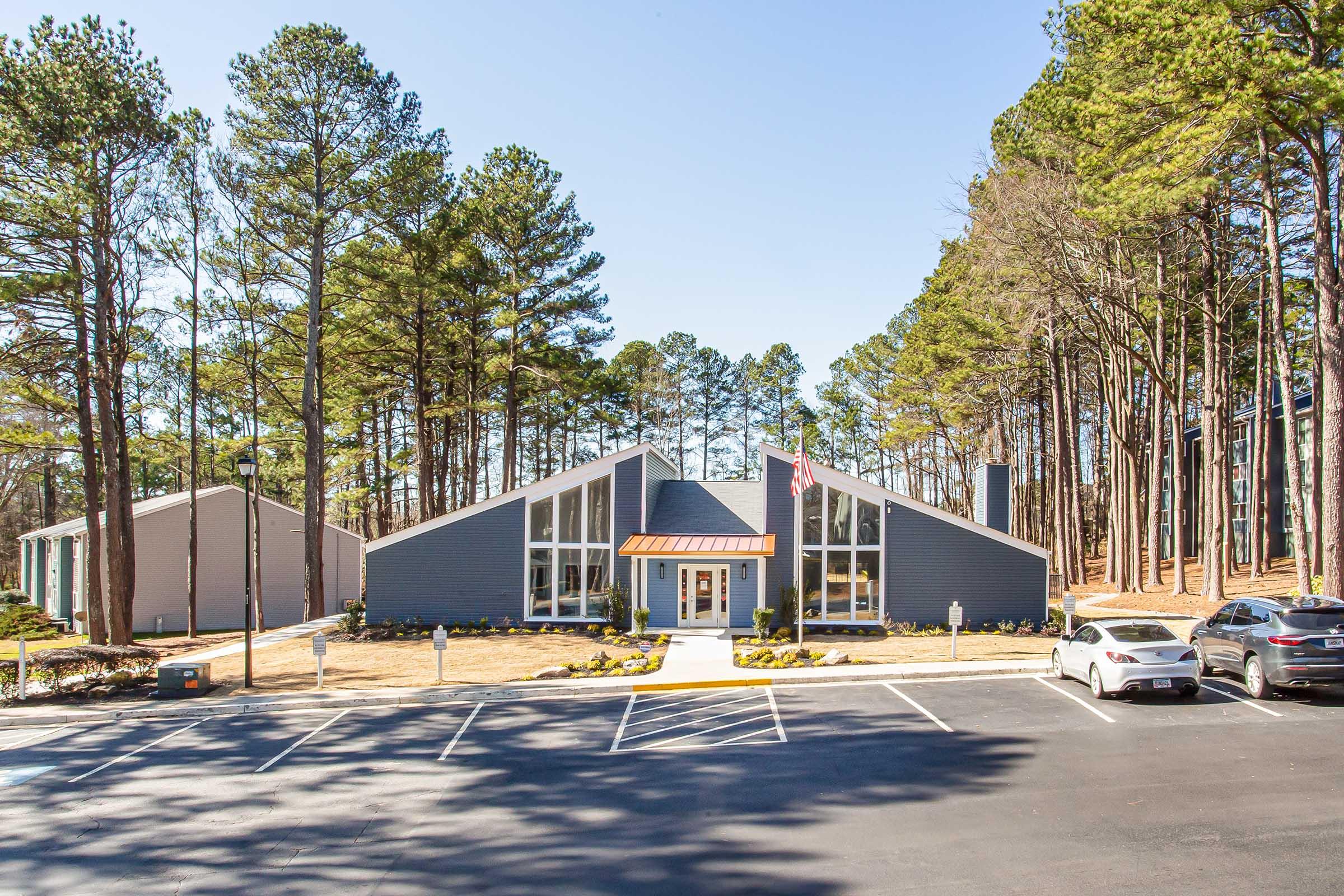 A modern building with a triangular design situated in a wooded area, surrounded by tall trees. The building features large windows and an entrance with steps leading up to it. There are several parked cars in a blacktop lot in front of the building, with a clear blue sky above.