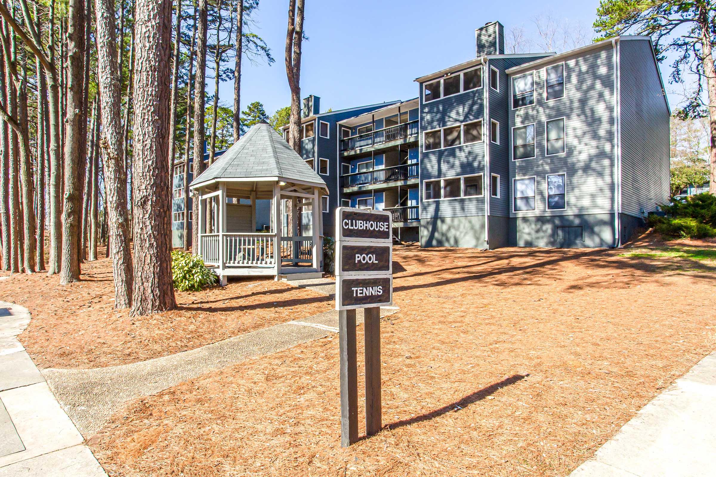 A landscaped area featuring a signpost with directions to a clubhouse, pool, and tennis court. In the background, a multi-story gray building is surrounded by tall trees, and a gazebo is visible nearby. The scene is well-lit and showcases a welcoming outdoor space.