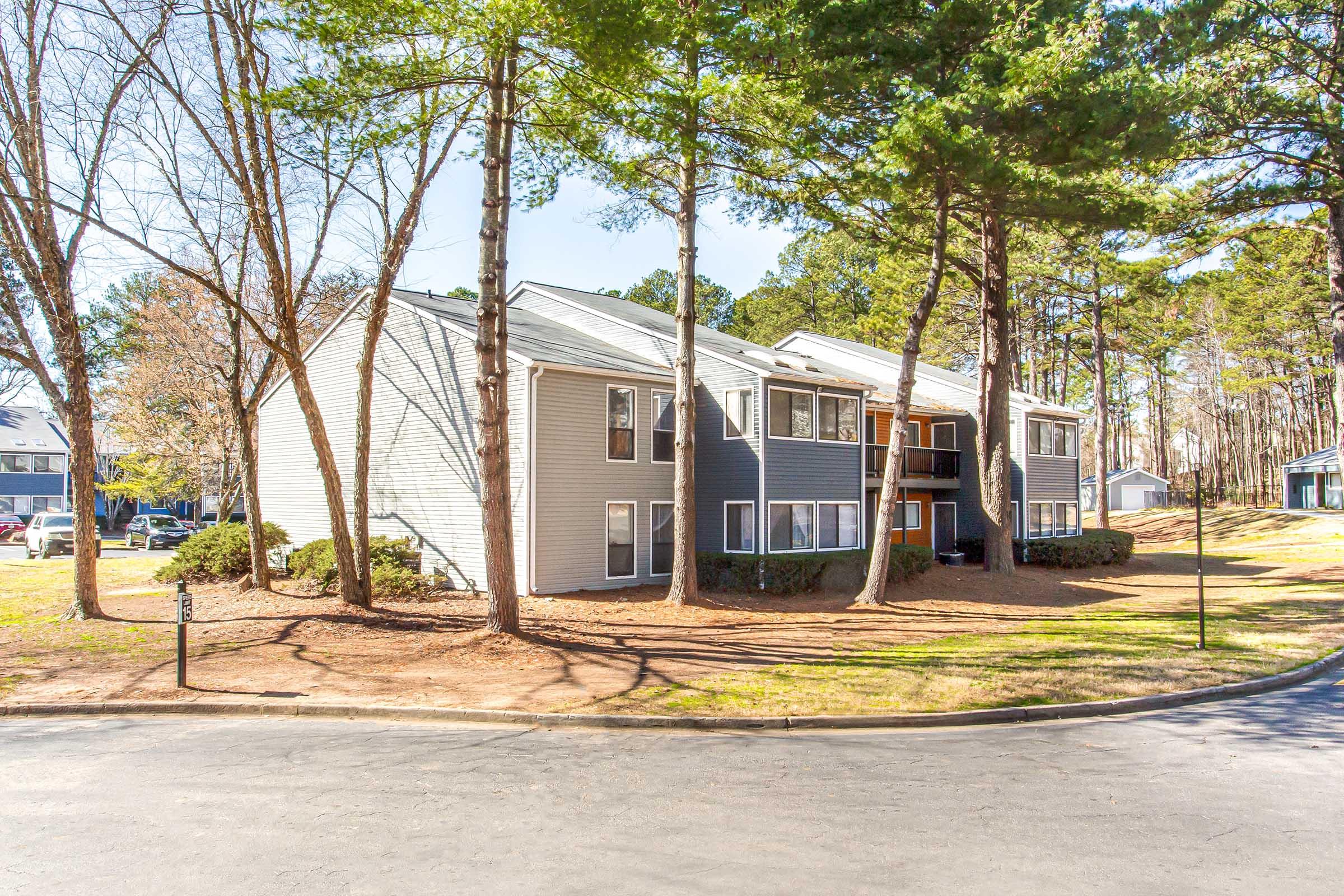 A residential apartment complex surrounded by tall pine trees, featuring multiple stories and a gray exterior. The building is situated at the corner of a street with a landscaped area in front, showcasing a serene, wooded environment typical for suburban living.