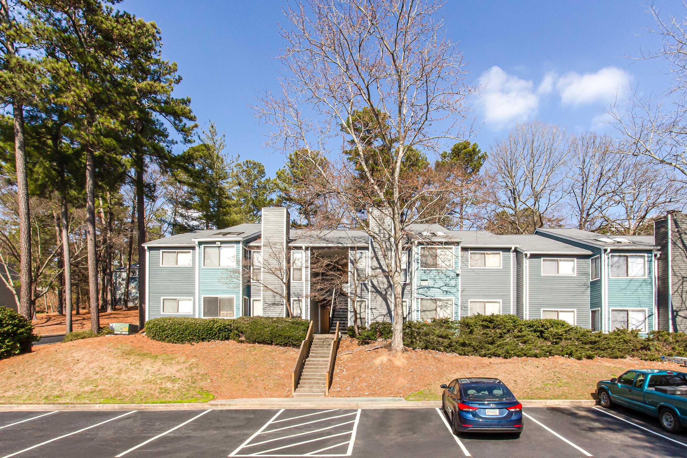 A two-story apartment building with a light blue exterior situated among trees. The area features a sloped lawn with shrubs and a parking lot in the foreground, where a blue car is parked. The sky is clear with a few clouds.