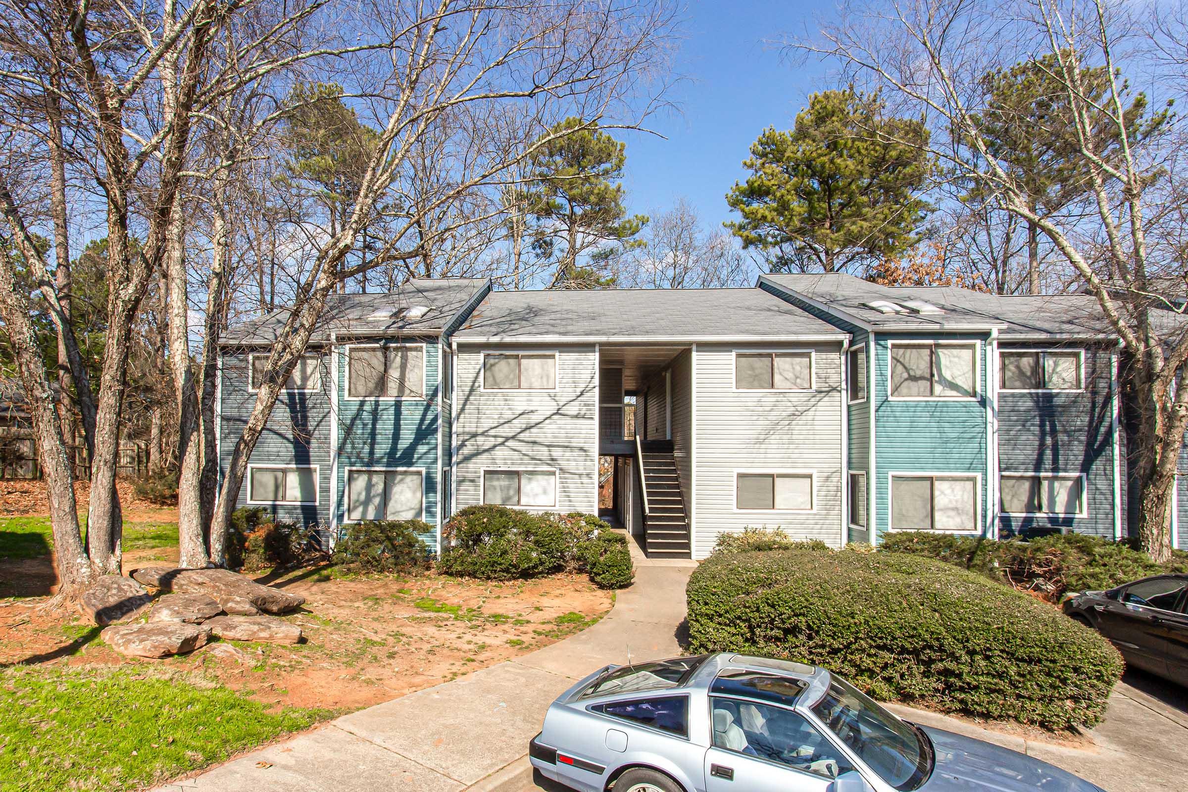 Exterior view of a two-story apartment building with a mix of blue and gray siding. Surrounded by trees and shrubbery, the building features multiple windows and a central entrance with stairs leading to the second floor. A car is parked in front on a paved path.