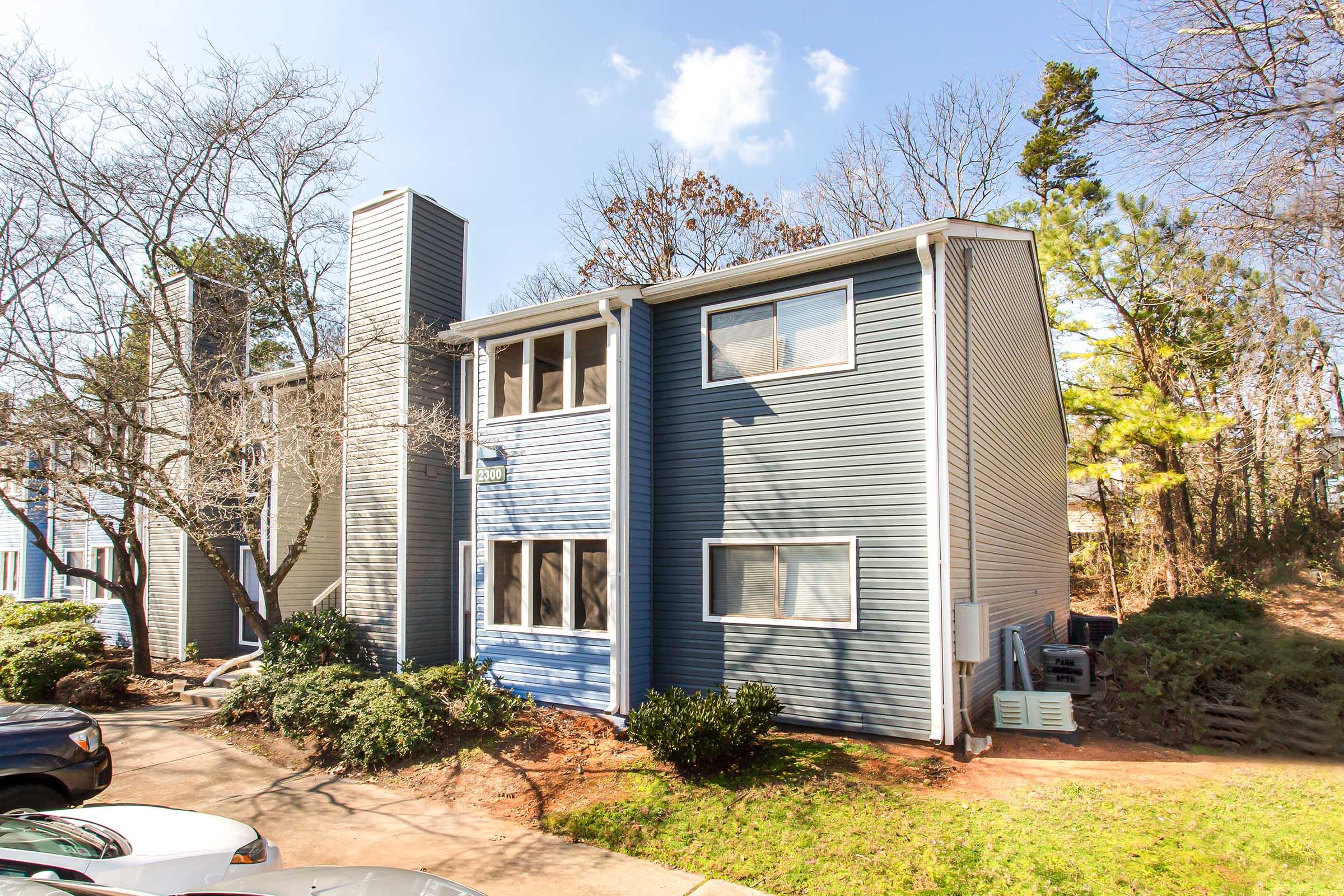 A two-story residential building featuring a blue and gray exterior, surrounded by trees and shrubs. The building has multiple windows and is situated next to a driveway with parked cars. The sky is clear with a few clouds, indicating a sunny day.