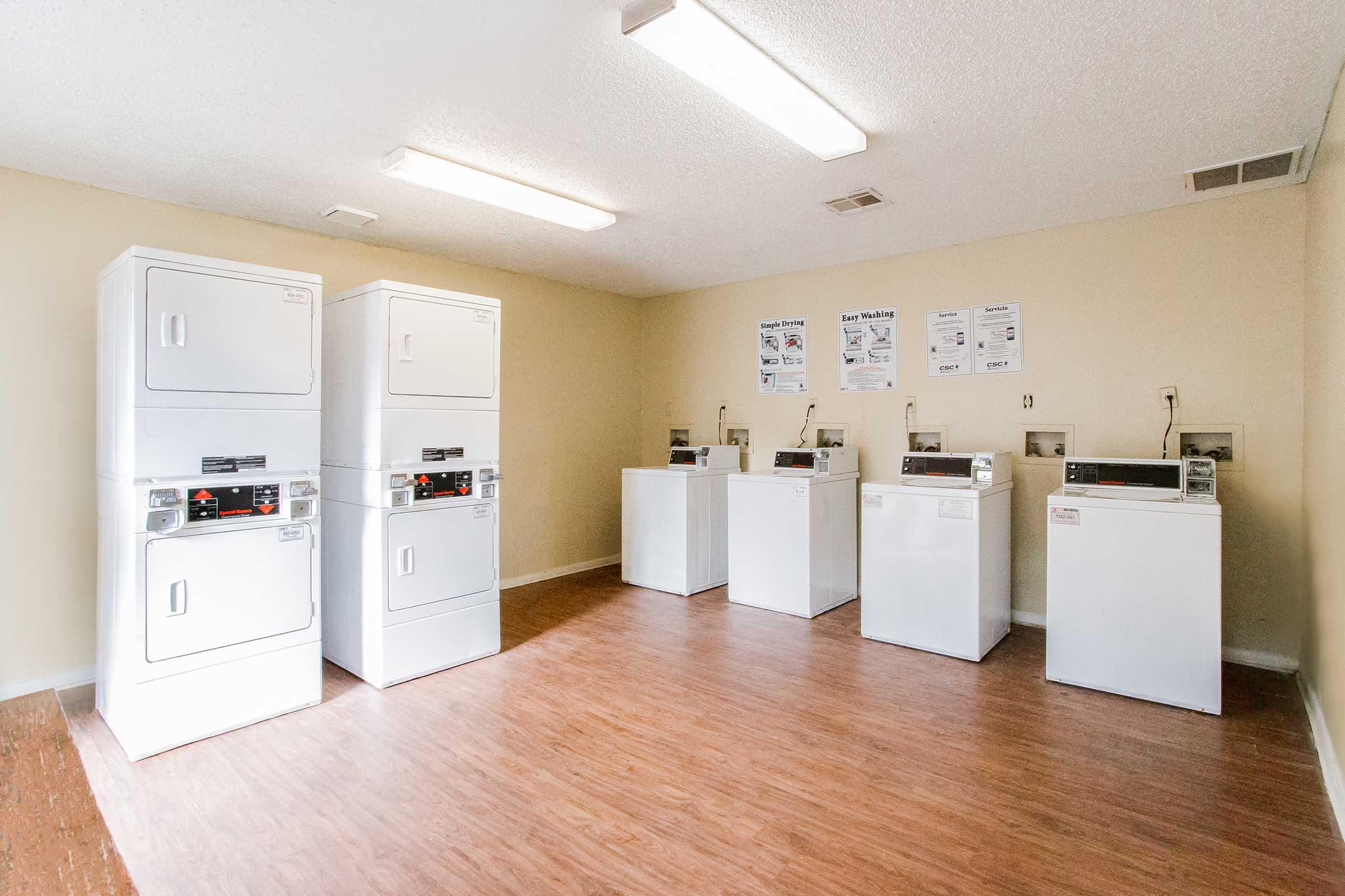 A clean laundry room featuring multiple white commercial washing machines and dryers. The floor is made of wood-like laminate, and there are fluorescent lights overhead. On the walls, there are informational posters about laundry usage and policies.