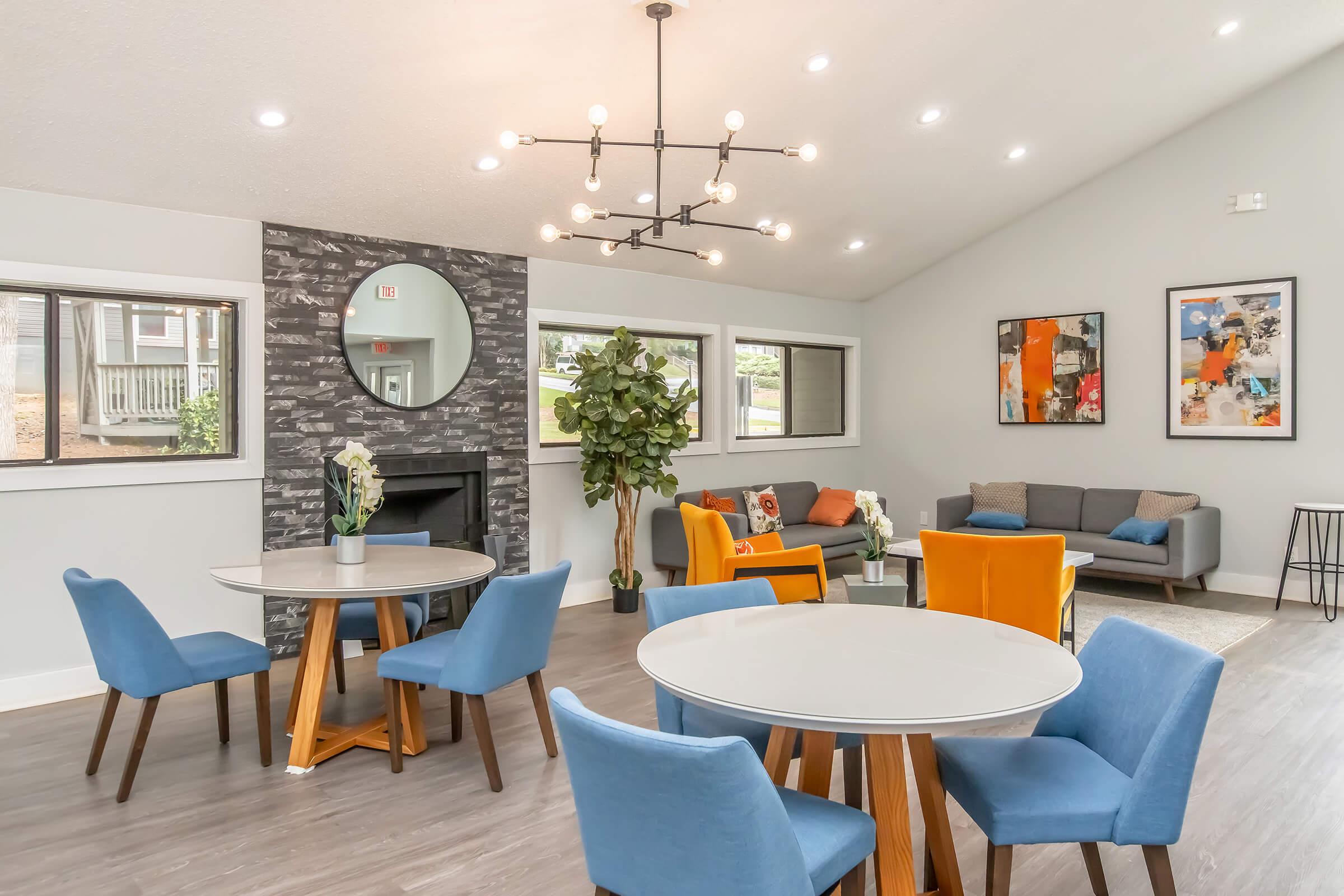 A modern living space featuring a round dining table with four blue chairs, a cozy seating area with gray and orange sofas, a stylish wall mirror, and decorative plants. Natural light floods the room through large windows, creating a welcoming atmosphere with contemporary decor.