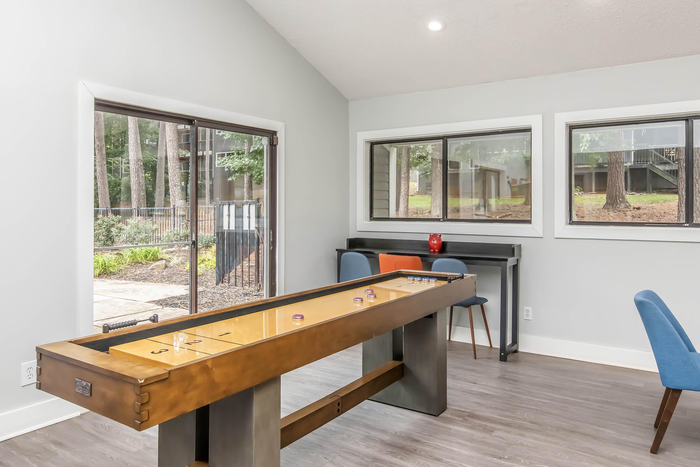 A spacious interior featuring a game table in the foreground, surrounded by blue chairs. Large windows provide a view of a wooded area outside, allowing natural light to brighten the room. The walls are painted a soft gray, contributing to a modern and inviting atmosphere.