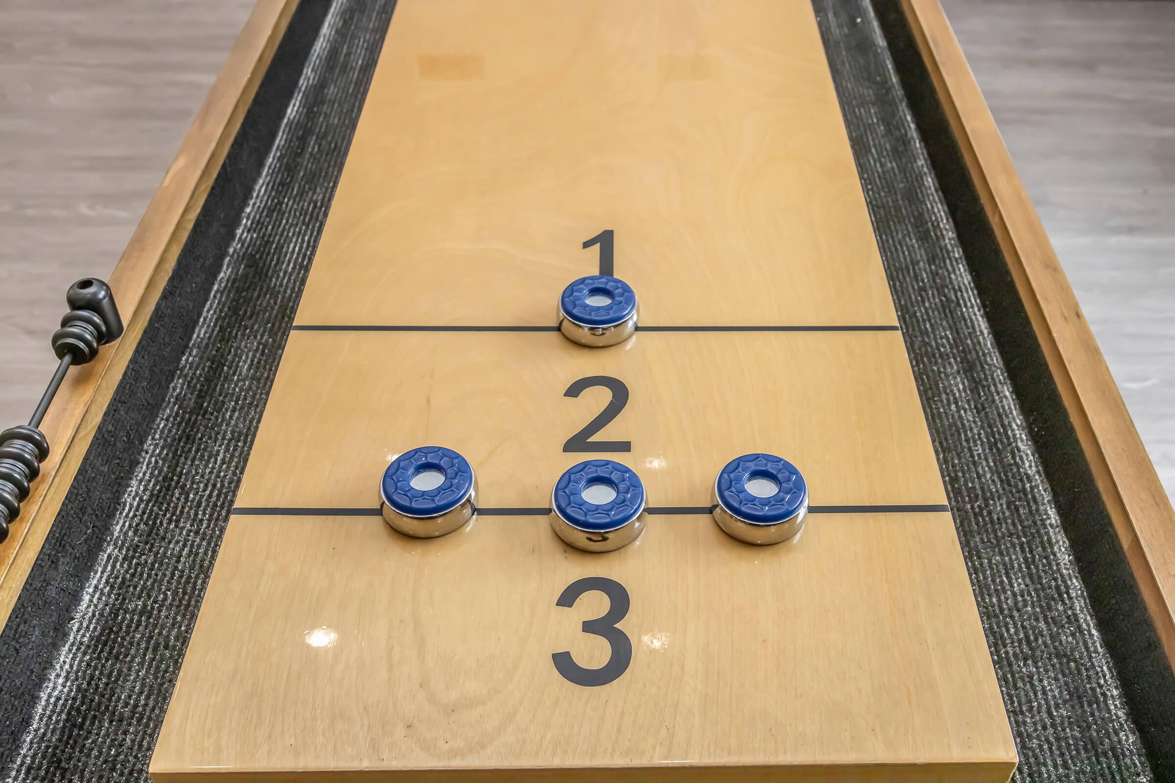 Close-up of a shuffleboard table featuring three blue pucks positioned on marked sections numbered 1, 2, and 3. The wooden surface is smooth, and a dark fabric runs along the sides, highlighting the game area. The scene captures a moment in a casual game setting.