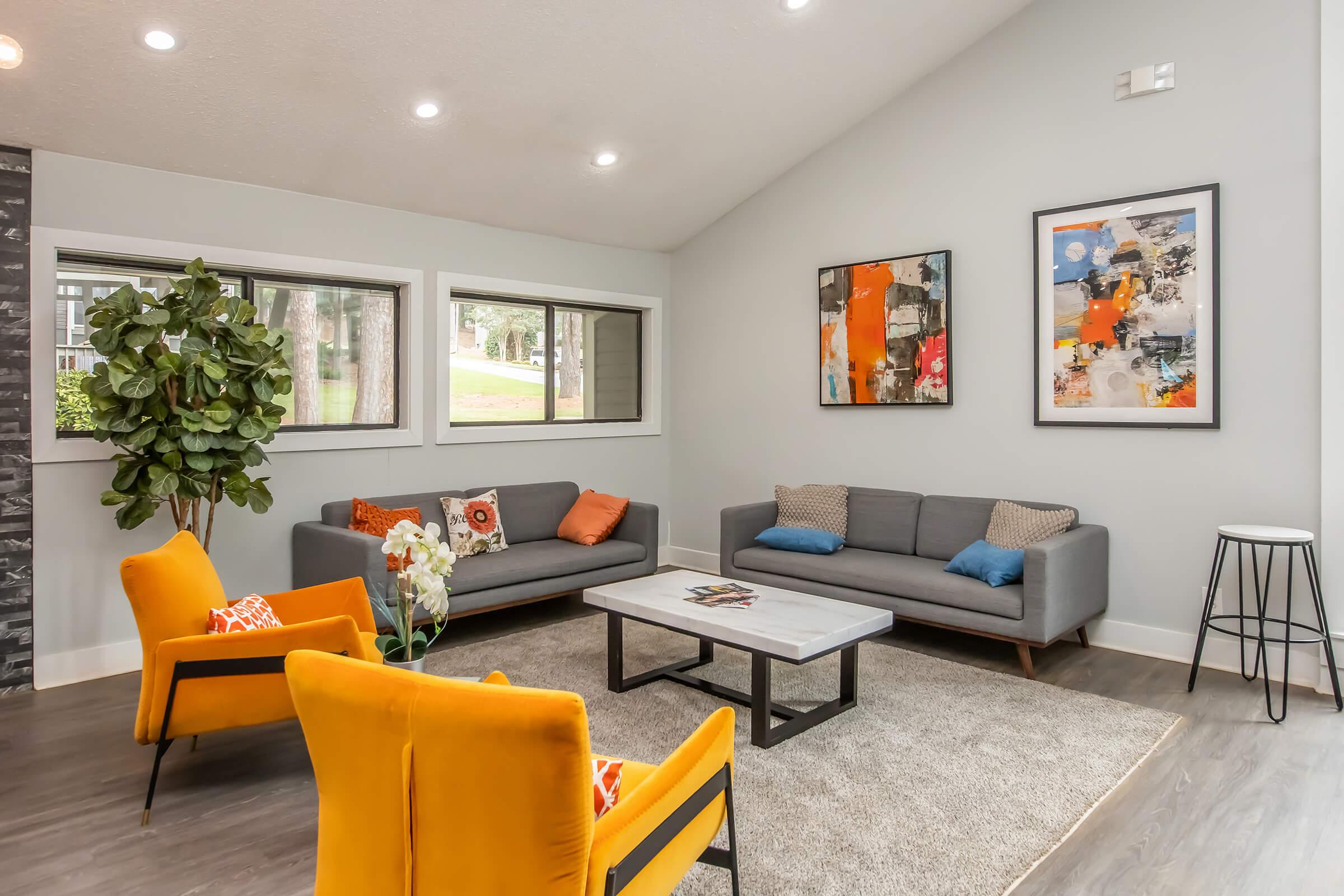 A modern living room featuring two gray sofas with colorful pillows, a coffee table, and two bright yellow accent chairs. A large area rug covers the floor, and there are two abstract art pieces on the walls. Natural light streams in through windows, complemented by a potted plant in the corner.