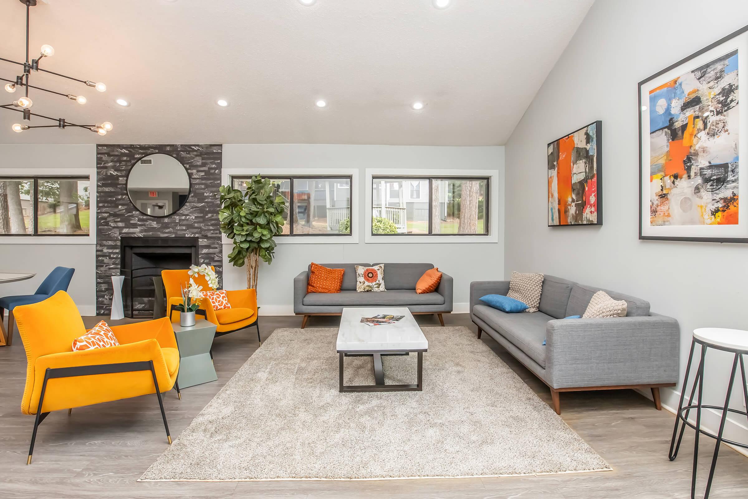 A modern living room featuring a gray sofa, vibrant orange armchairs, and a light gray coffee table on a large area rug. A decorative wall with artwork hangs above, and large windows provide natural light. A round mirror is positioned above the fireplace, enhancing the contemporary feel of the space.