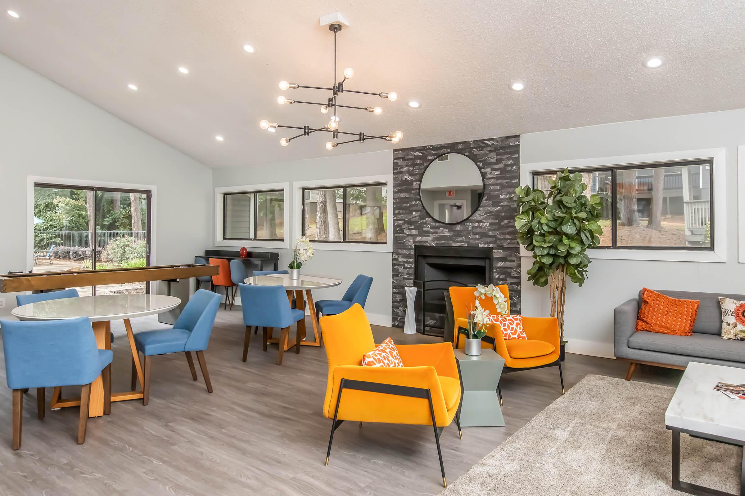 A modern living space featuring a cozy arrangement of bright orange and gray furniture, including chairs and a sofa. A sleek dining area with round tables and stylish light fixtures illuminates the room, complemented by large windows and a decorative plant. A fireplace adds warmth to the inviting atmosphere.