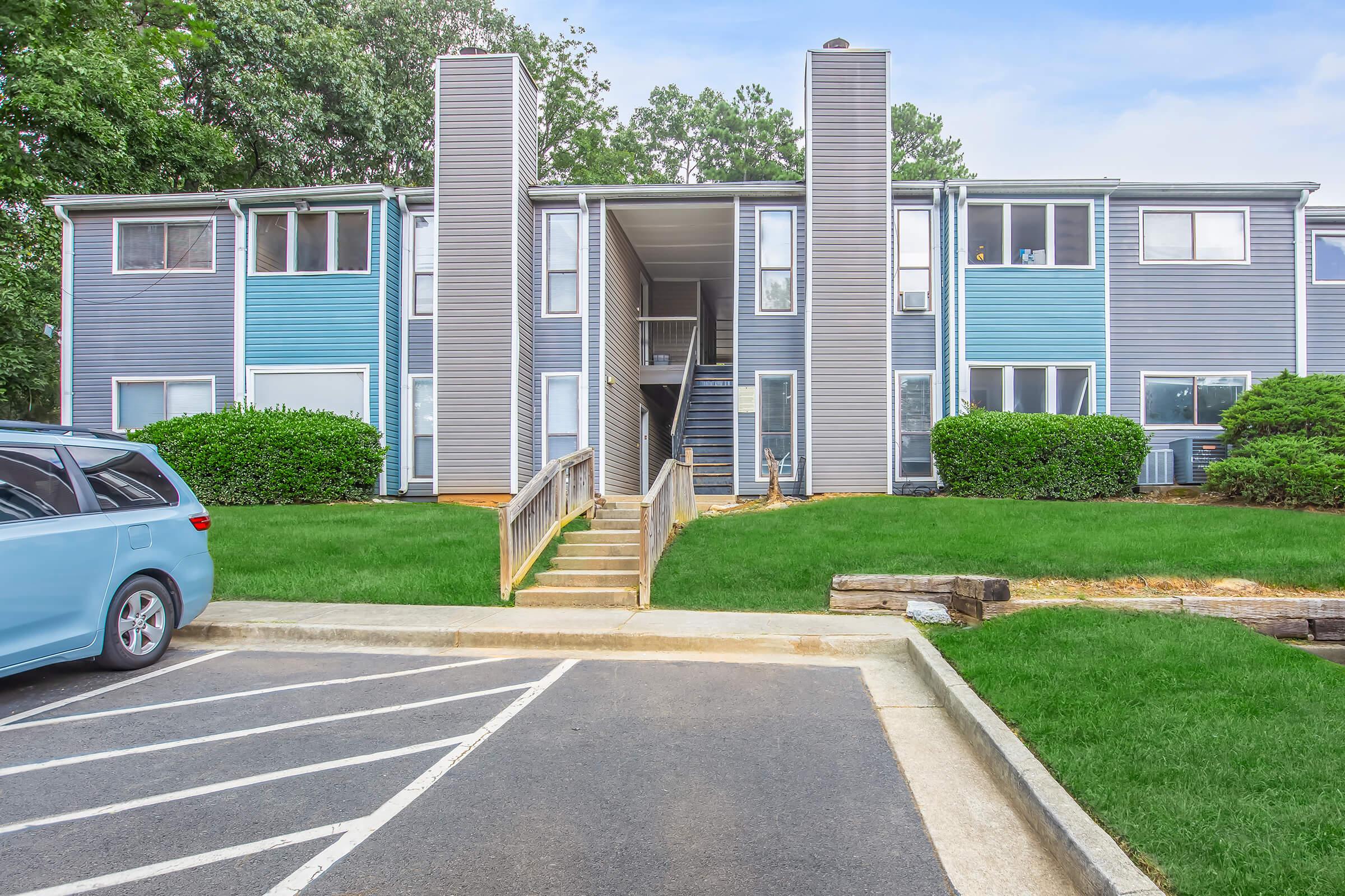Two-story apartment building with gray and blue siding, surrounded by green grass and shrubs. A set of wooden steps leads to the entrance, while a parking lot with a blue vehicle is visible in the foreground. The area is well-maintained under a clear sky.