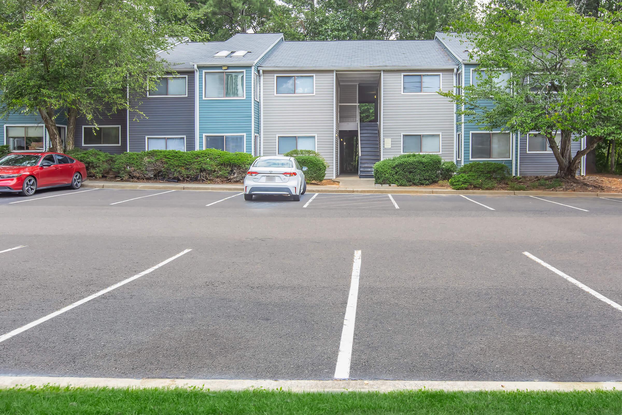 A view of an apartment complex with two buildings. The left building is light blue, while the right is gray. A red car is parked on the left, and a white car is parked in the center. The parking lot is mostly empty, surrounded by greenery and trees. The scene is set on a sunny day with clear weather.