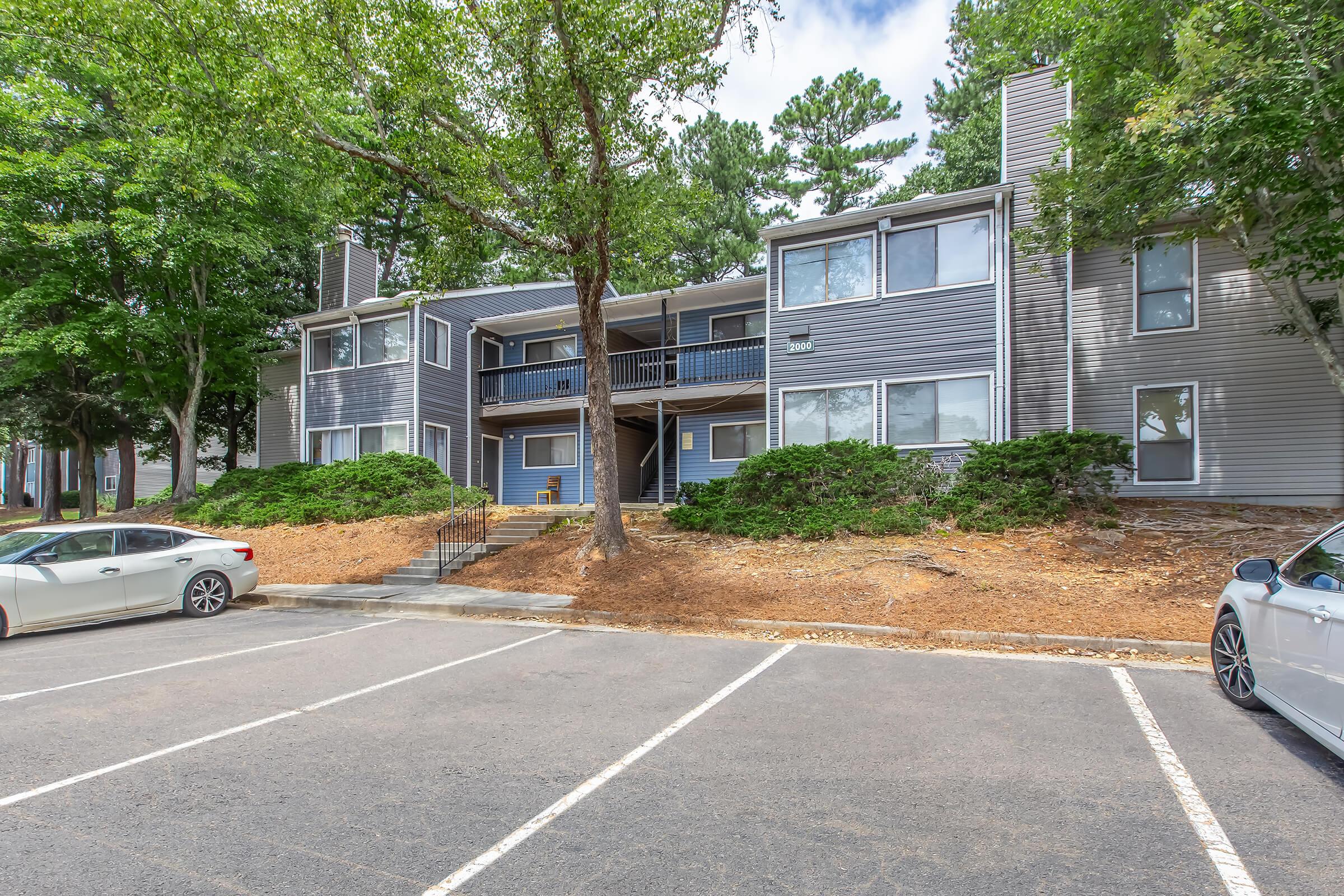 Exterior view of a two-story apartment building surrounded by trees and landscaping. The building features a mix of gray siding and balconies. A parking lot with parked cars is visible in the foreground, and the sky is partly cloudy.