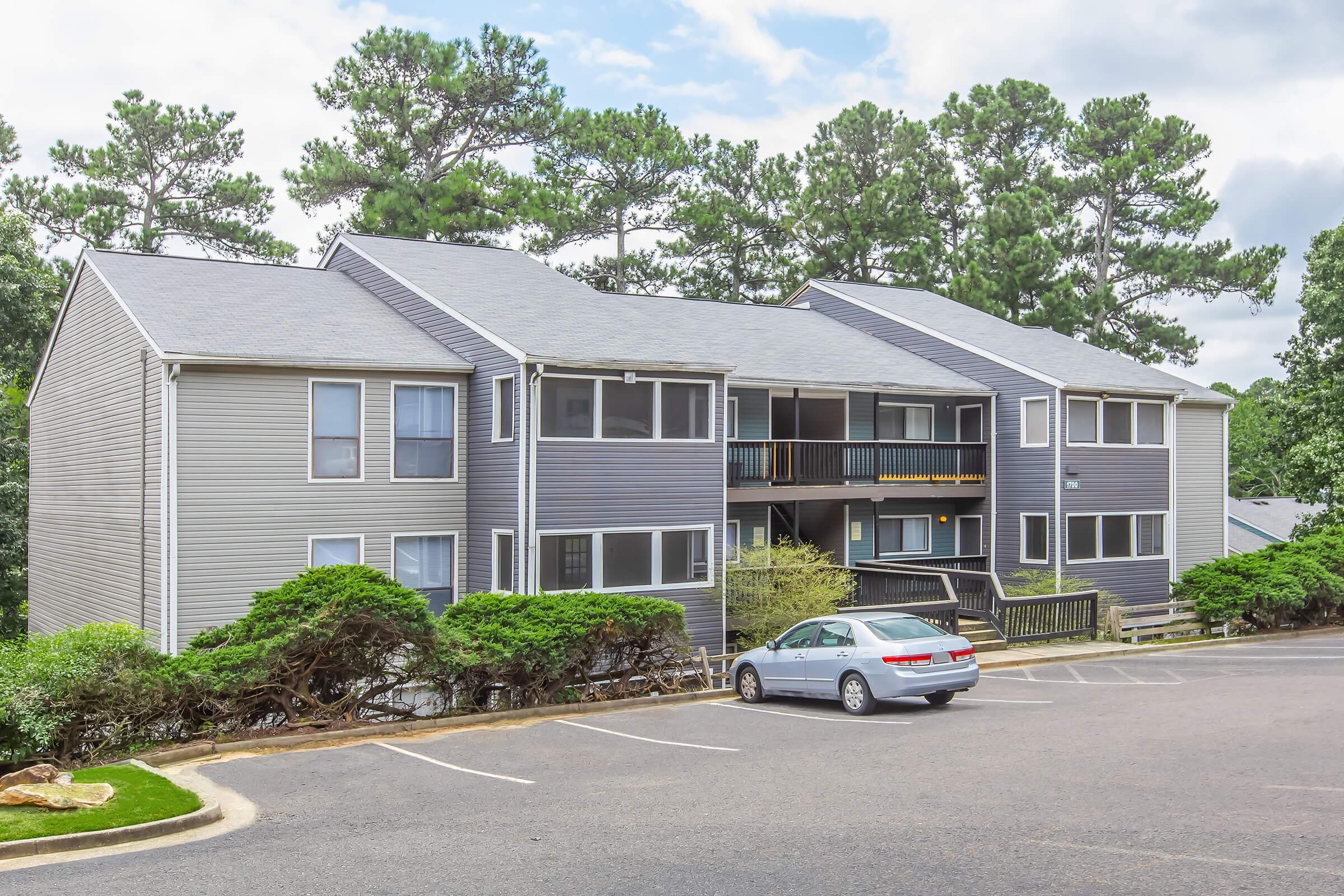 A multi-unit apartment building featuring gray siding and a front porch area. The structure has two levels with balconies. In front, there is a parking lot with a white car parked. Lush green bushes line the front, and pine trees are visible in the background under a partly cloudy sky.