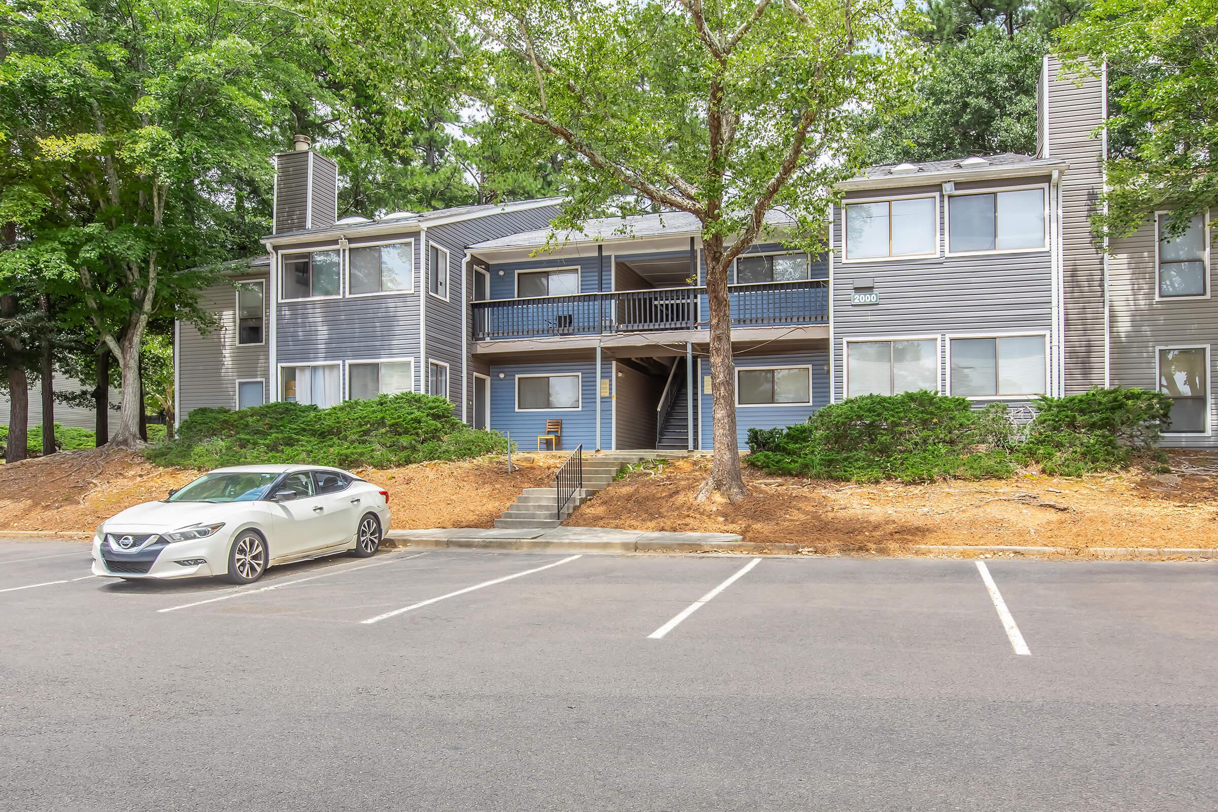 A two-story apartment building with grey siding and multiple windows, surrounded by green trees. A small staircase leads to the entrance, and there is a white car parked in the foreground on a paved parking lot. The area features landscaped shrubs and mulch.